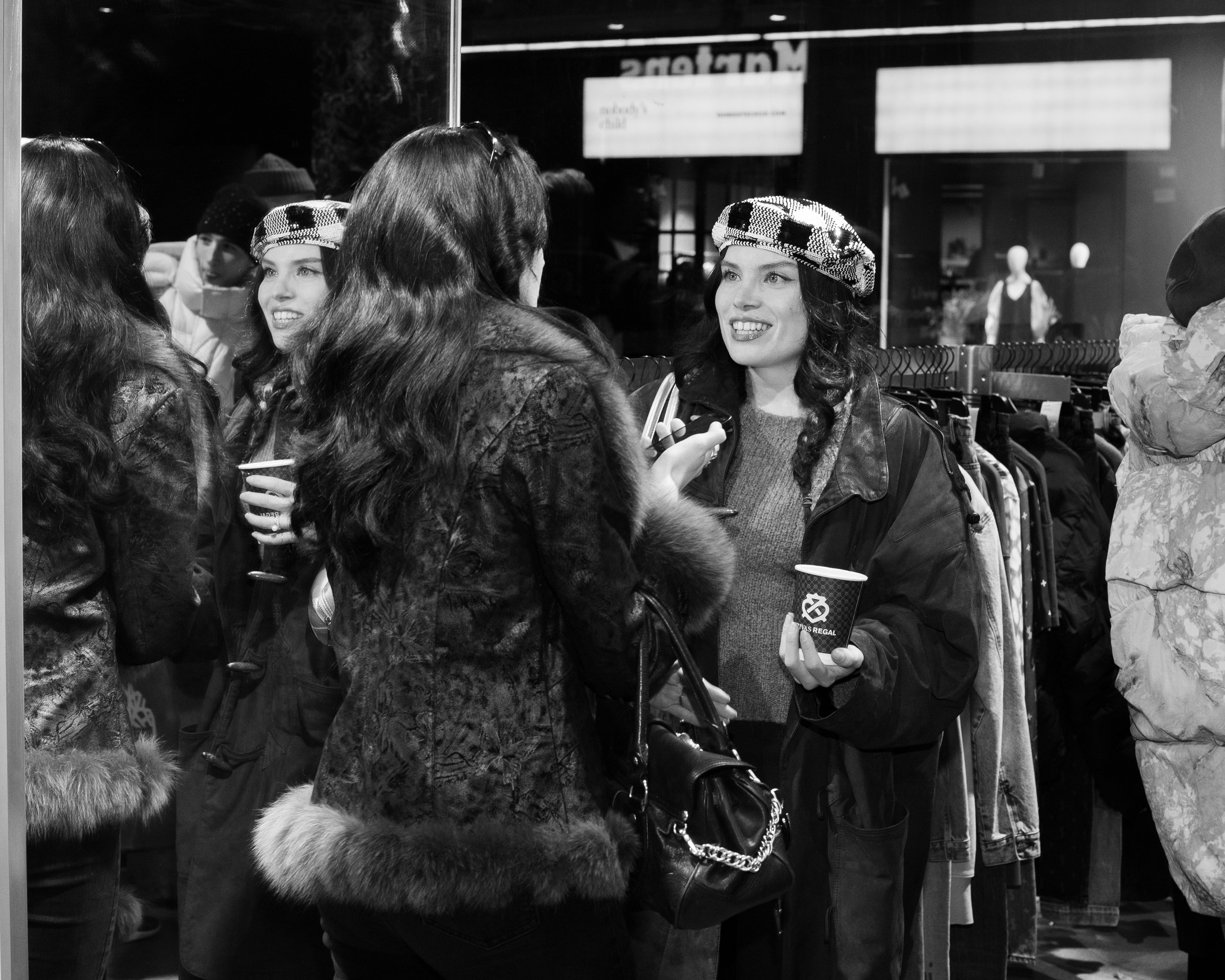 People talking inside a store, two women in the center are facing each other and smiling, one holds a drink, the other has a handbag. Others are visible in the background.