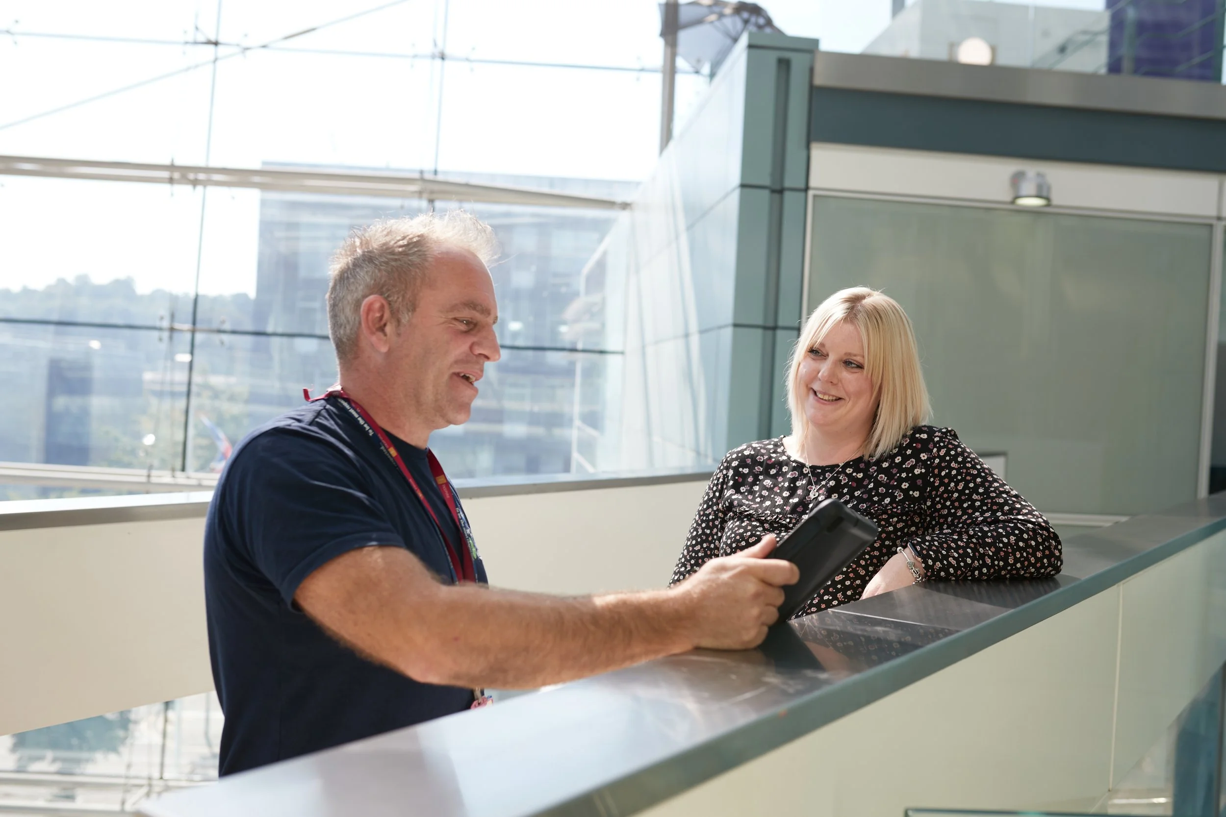 Two people, a man and a woman, are smiling and talking to each other in a modern indoor space with large glass windows and a city view. The man is holding a smartphone.