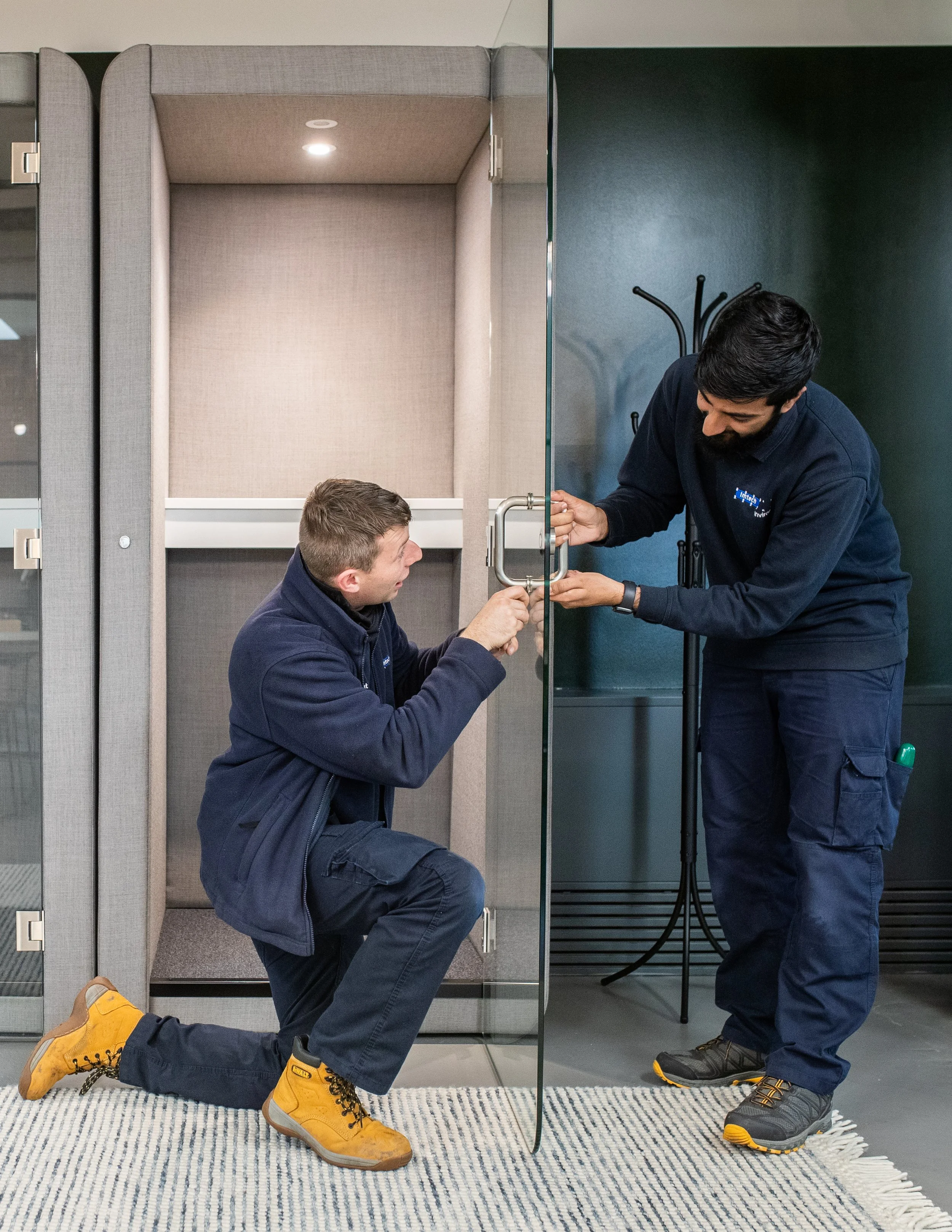 Two men installing a glass door handle in an office or commercial space, with one kneeling and holding the handle while the other is screwing it in.