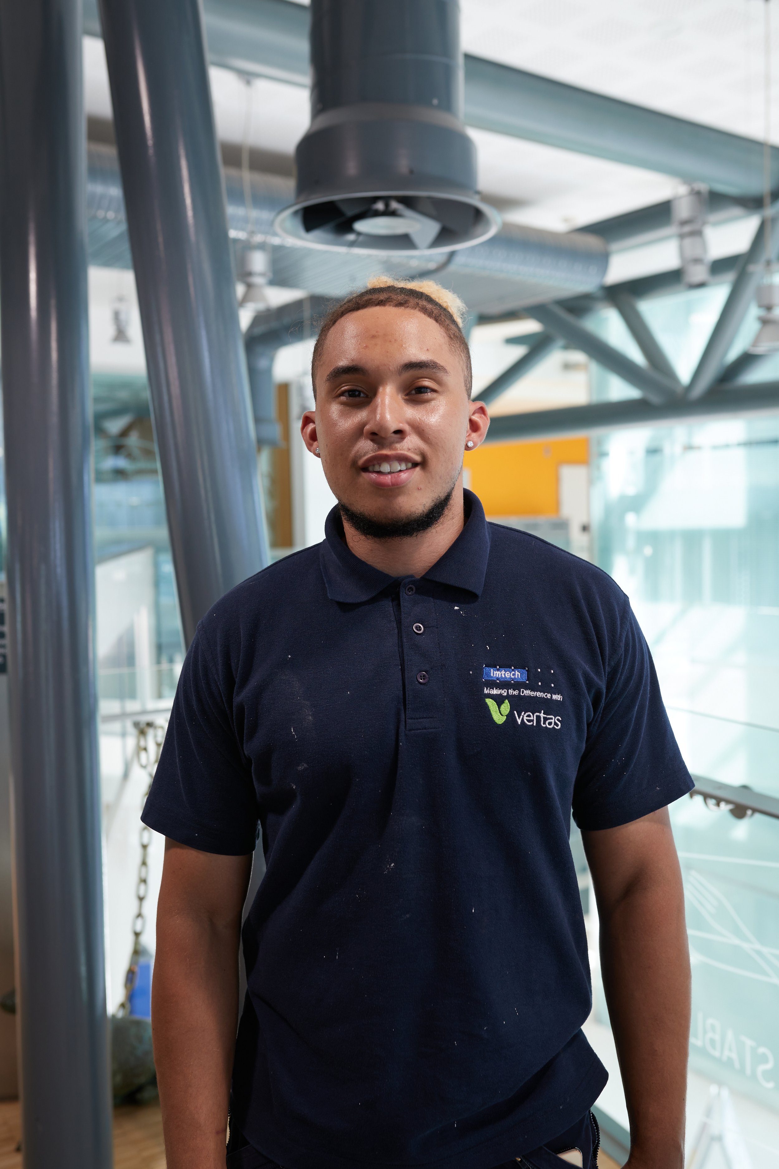 Young man wearing a navy polo shirt with logos standing indoors in a modern building.