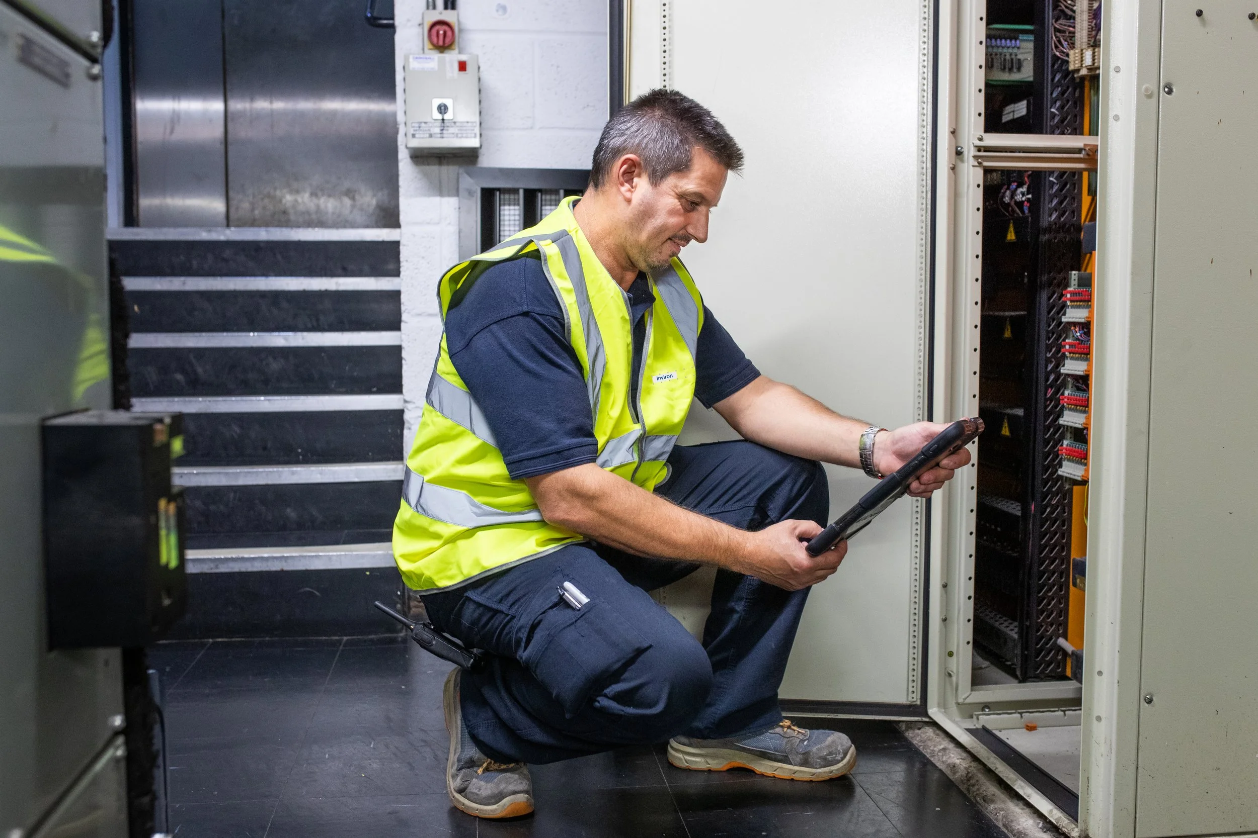 A technician in a yellow safety vest kneeling and inspecting an electrical cabinet with a tablet in hand.