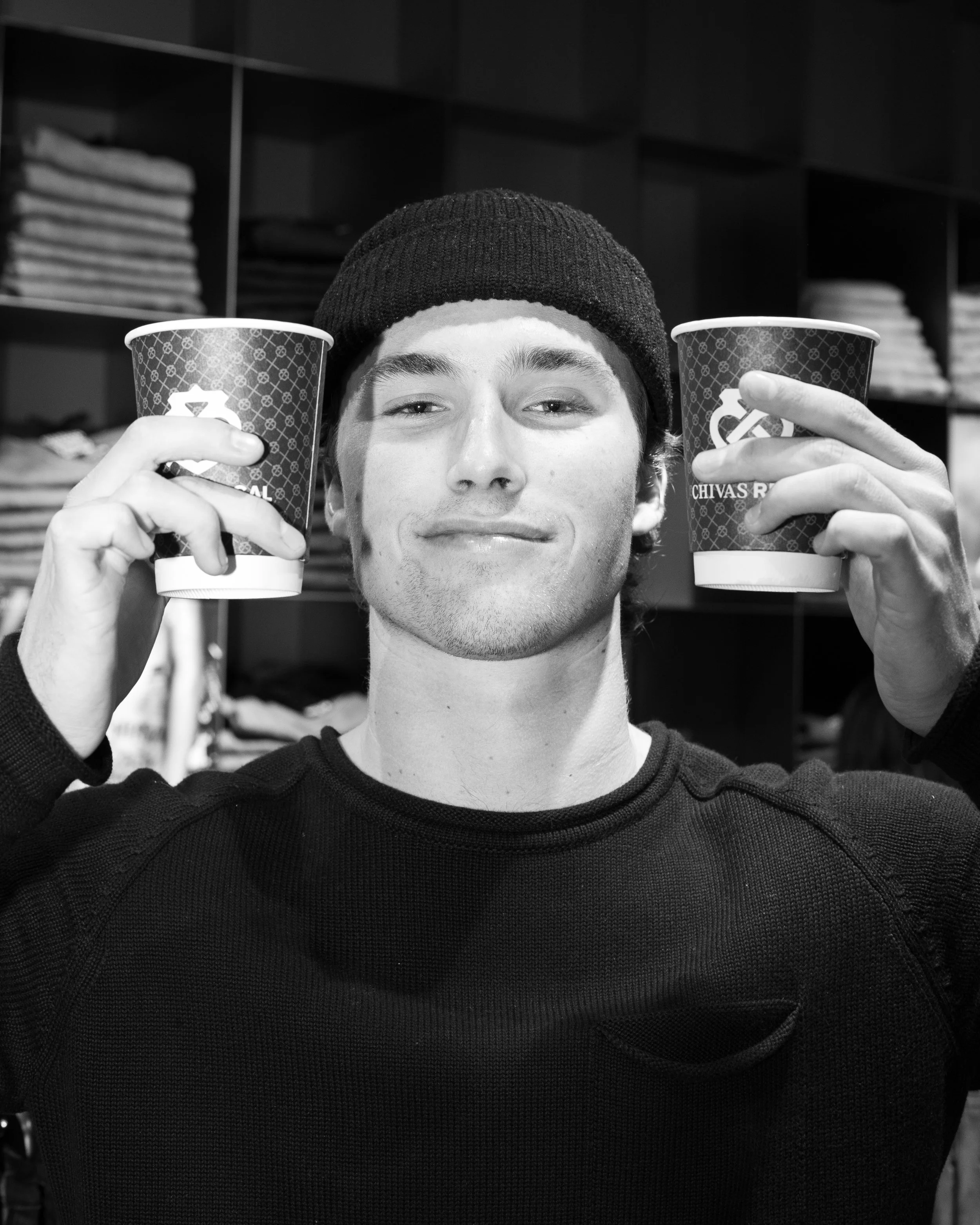 A young man with a beanie and a black shirt smiling and holding two cups, one in each hand, in front of a background with shelves of folded clothes.