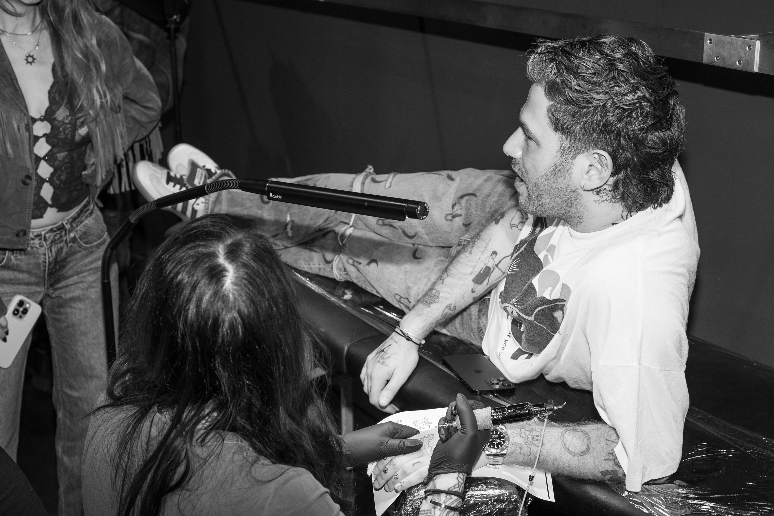 A man with tattoos on his arm and long hair is sitting at a table while getting a tattoo. A woman wearing gloves is tattooing his hand. There are people standing nearby, and one person is holding a phone.