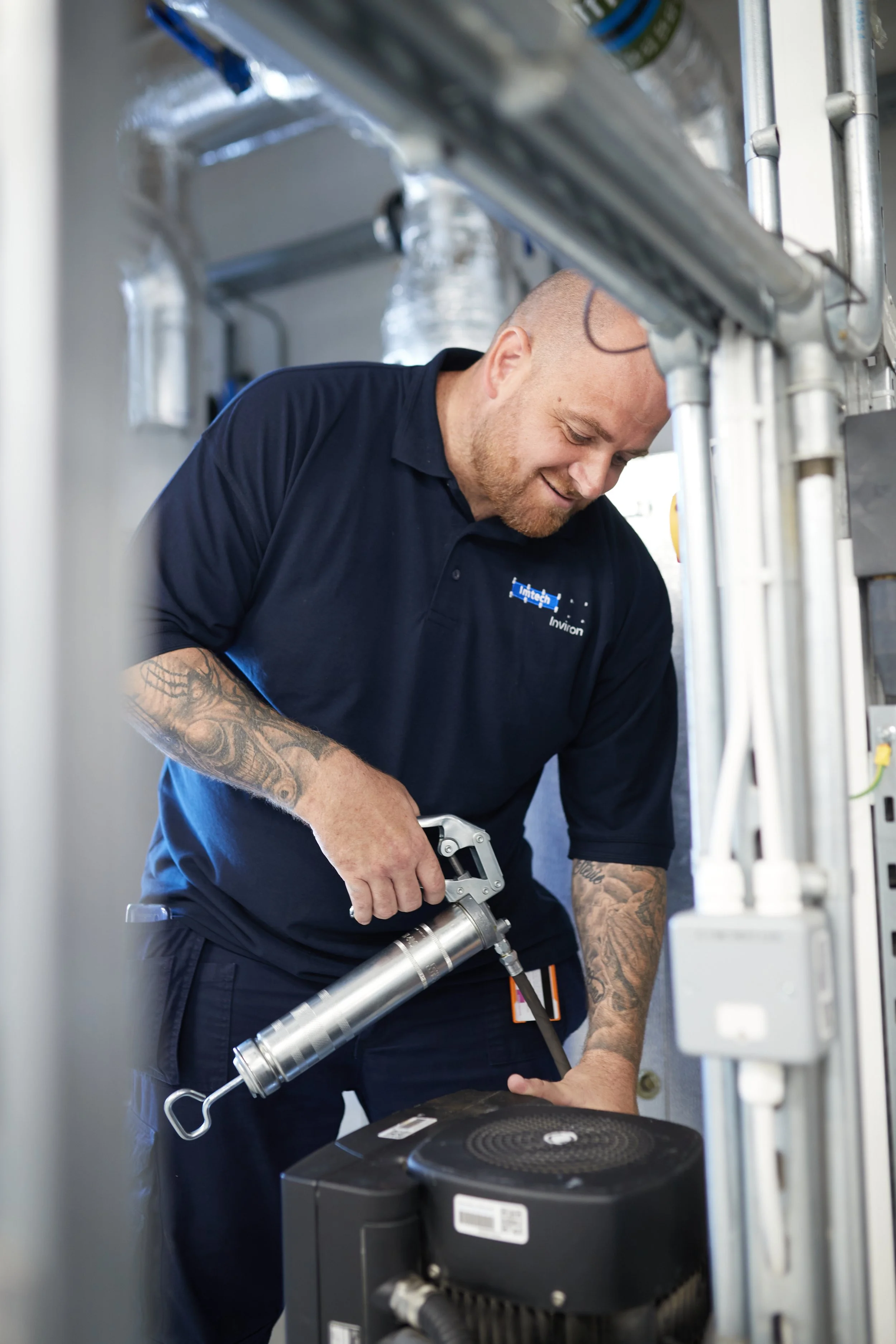 A technician with tattoos on his arms wearing a navy blue polo shirt, smiling while working on equipment using a tool in a mechanical or industrial setting.