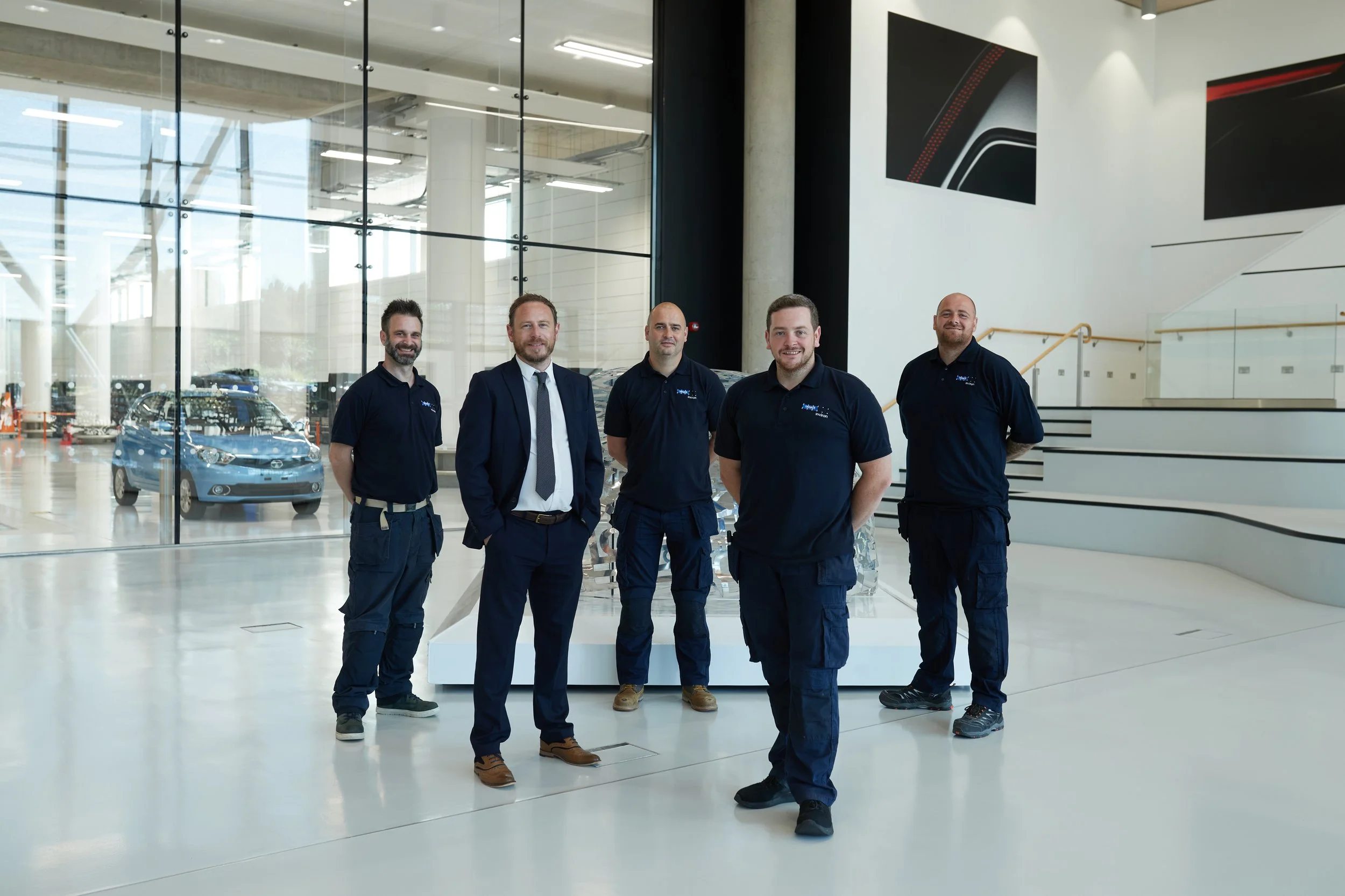 Group of five men standing inside a modern showroom, posing for a photo. The background features large glass windows and a car display.