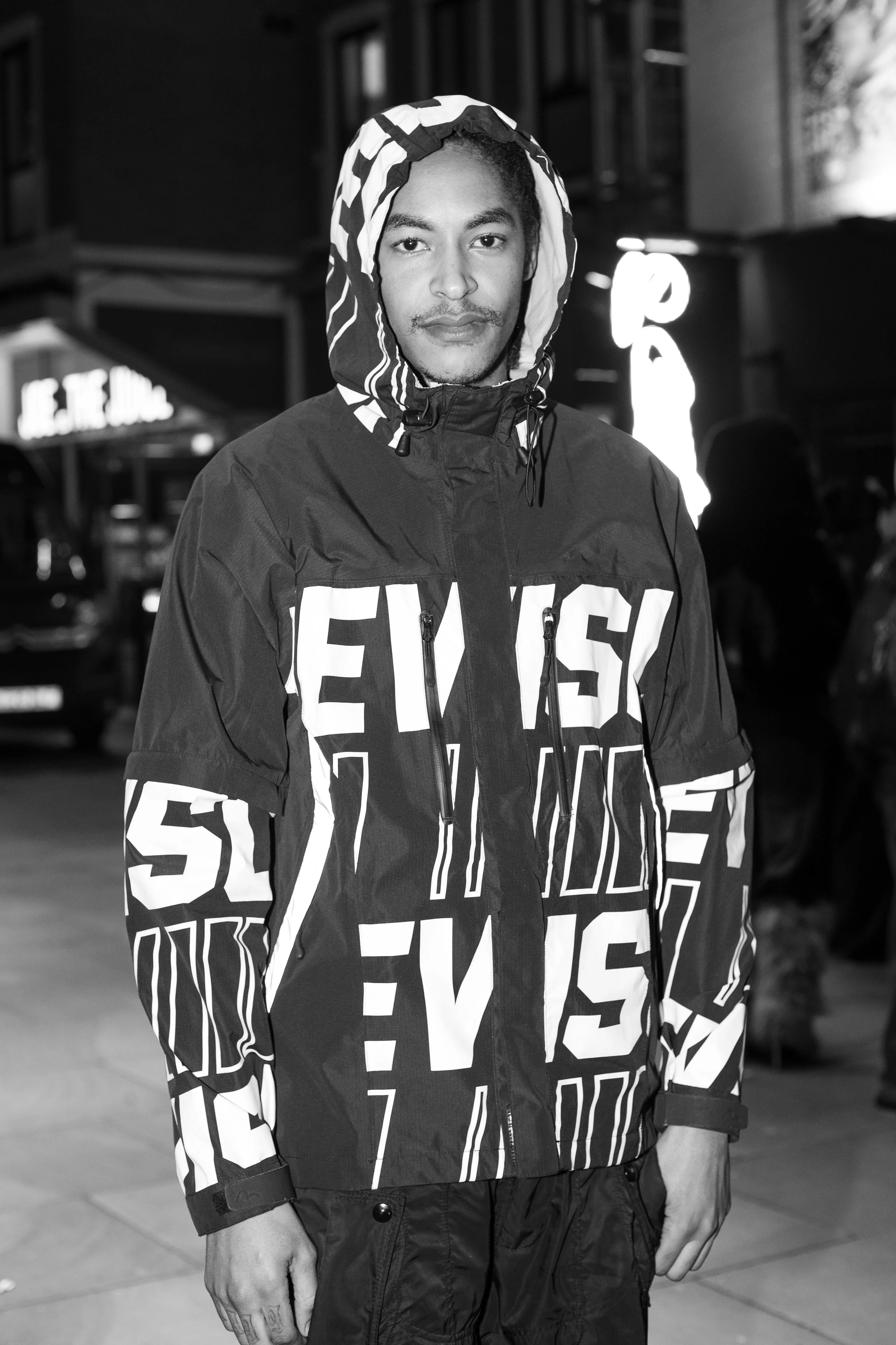 A young man with braided hair wearing a patterned hooded jacket, standing outdoors at night with city lights and buildings in the background.
