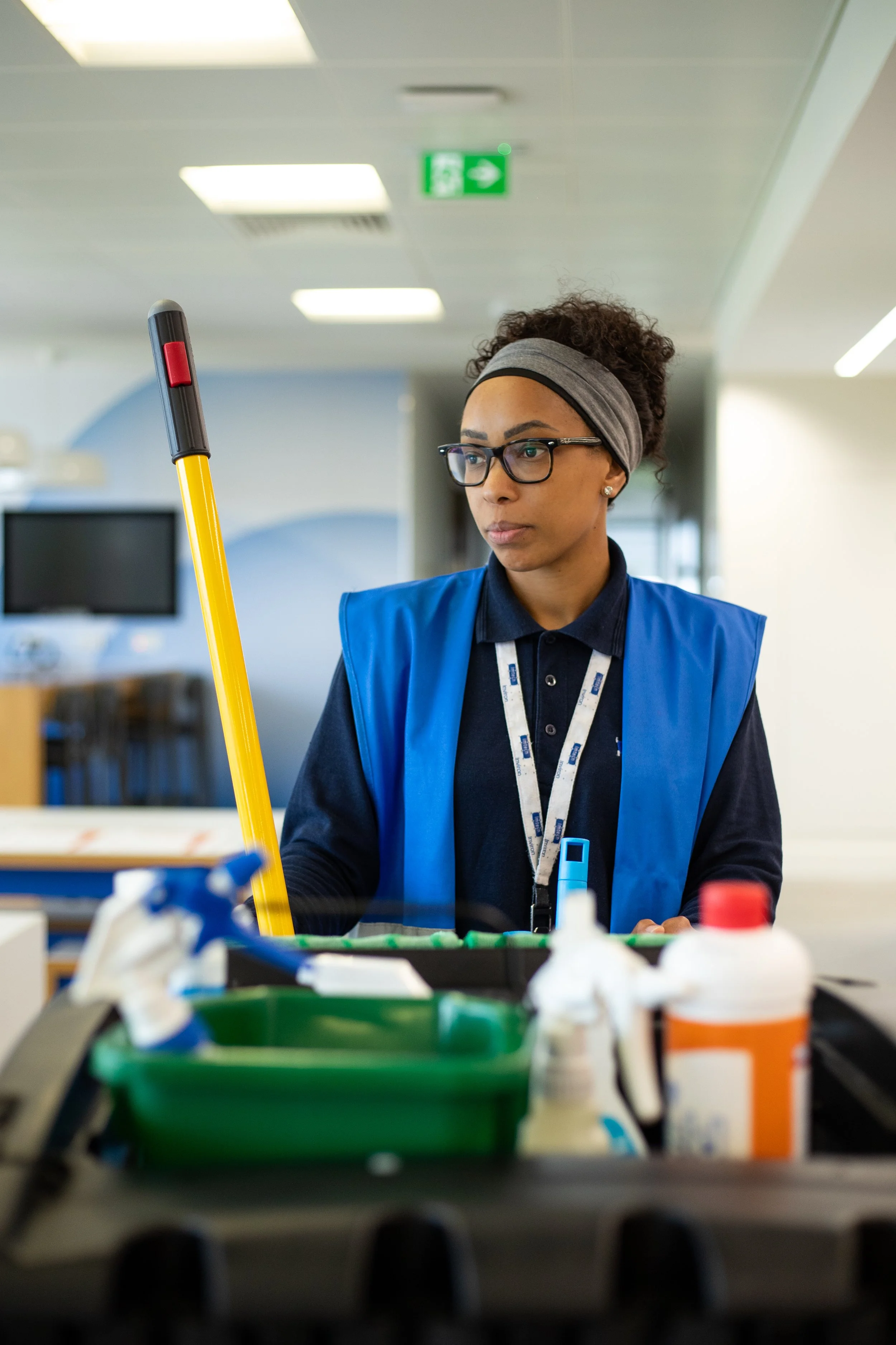 Female custodian wearing glasses and a blue vest, holding a mop, standing at a cleaning cart with cleaning supplies inside an indoor facility.