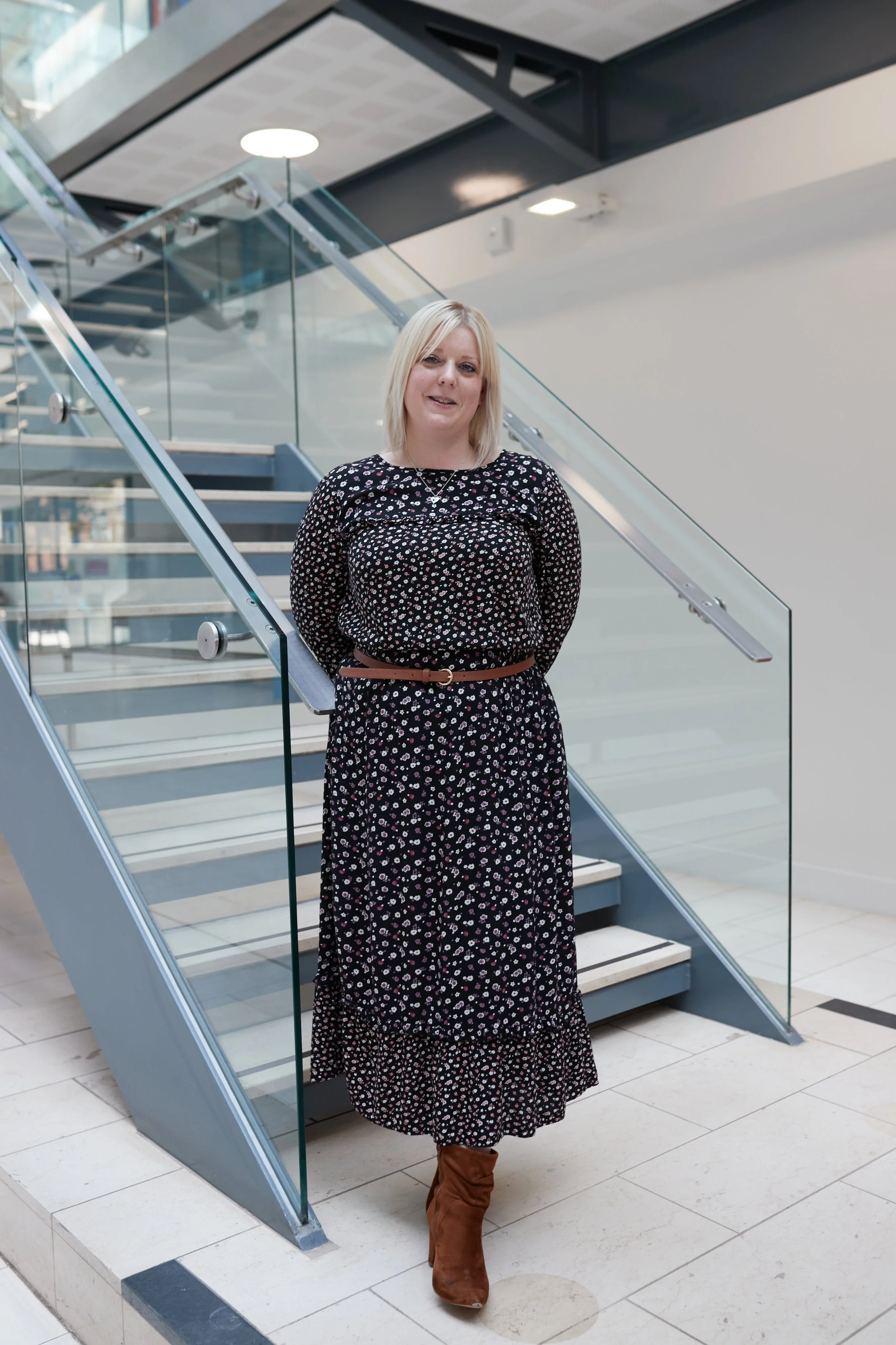 A woman with blonde hair standing on a staircase inside a modern building, wearing a long black floral dress, brown boots, and a brown belt.