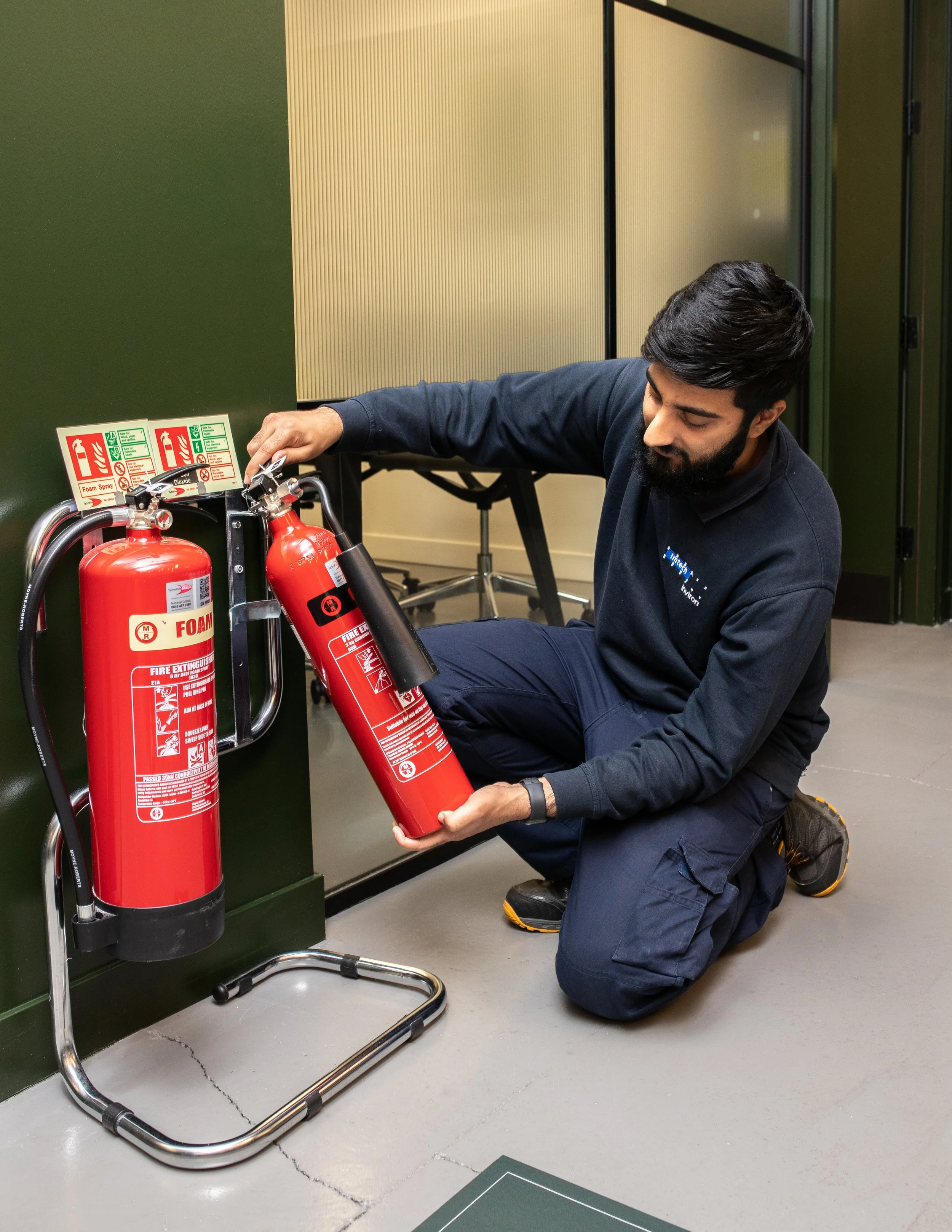 A man kneeling on the floor assembling or inspecting two red fire extinguishers mounted on a wall in an indoor setting.