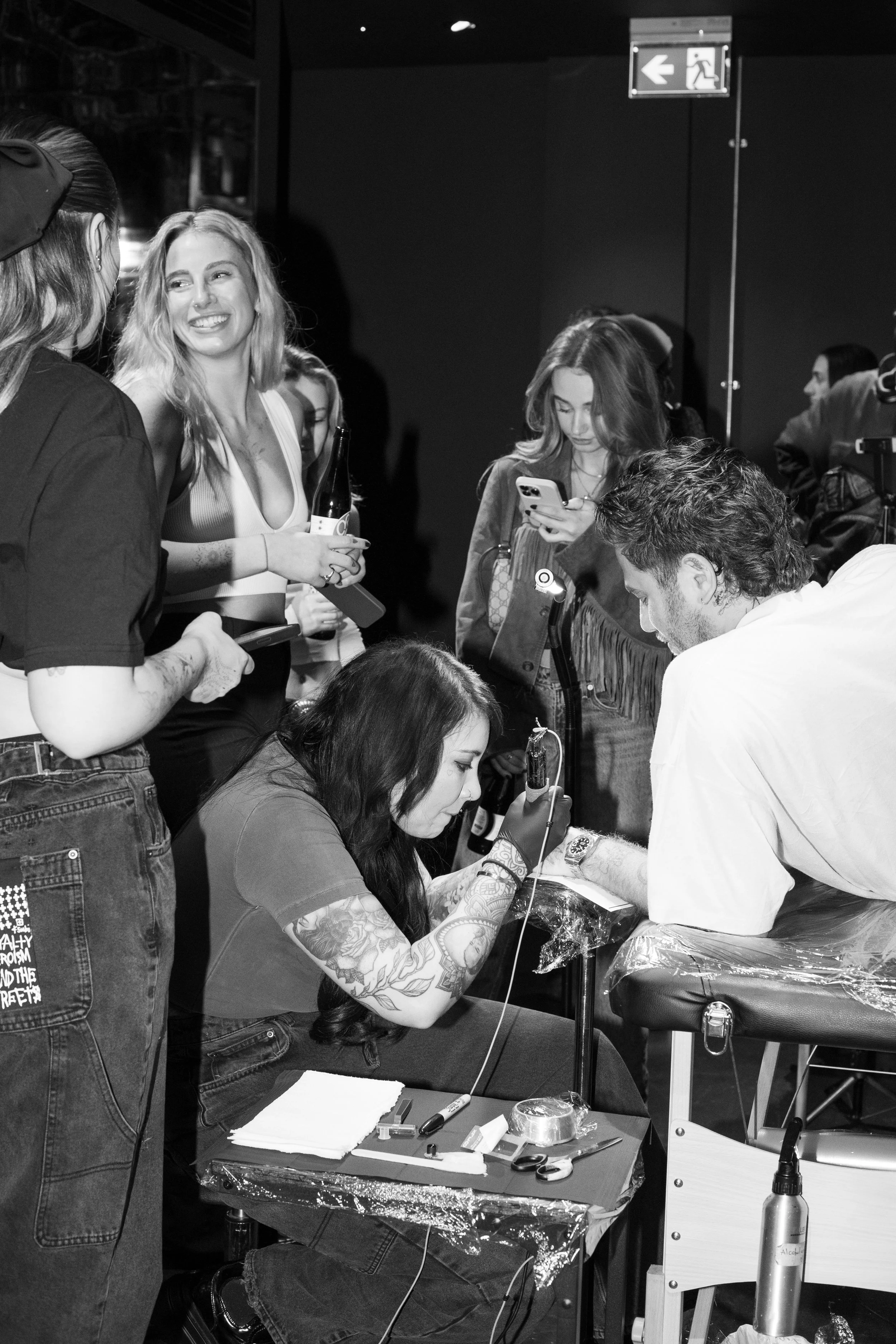 Black and white photo of a woman with long dark hair tattooed on her arms, laying on a table getting a tattoo. Several women and a man stand around watching, one woman smiling and holding a bottle, another woman looking at her phone, and the man tatt
