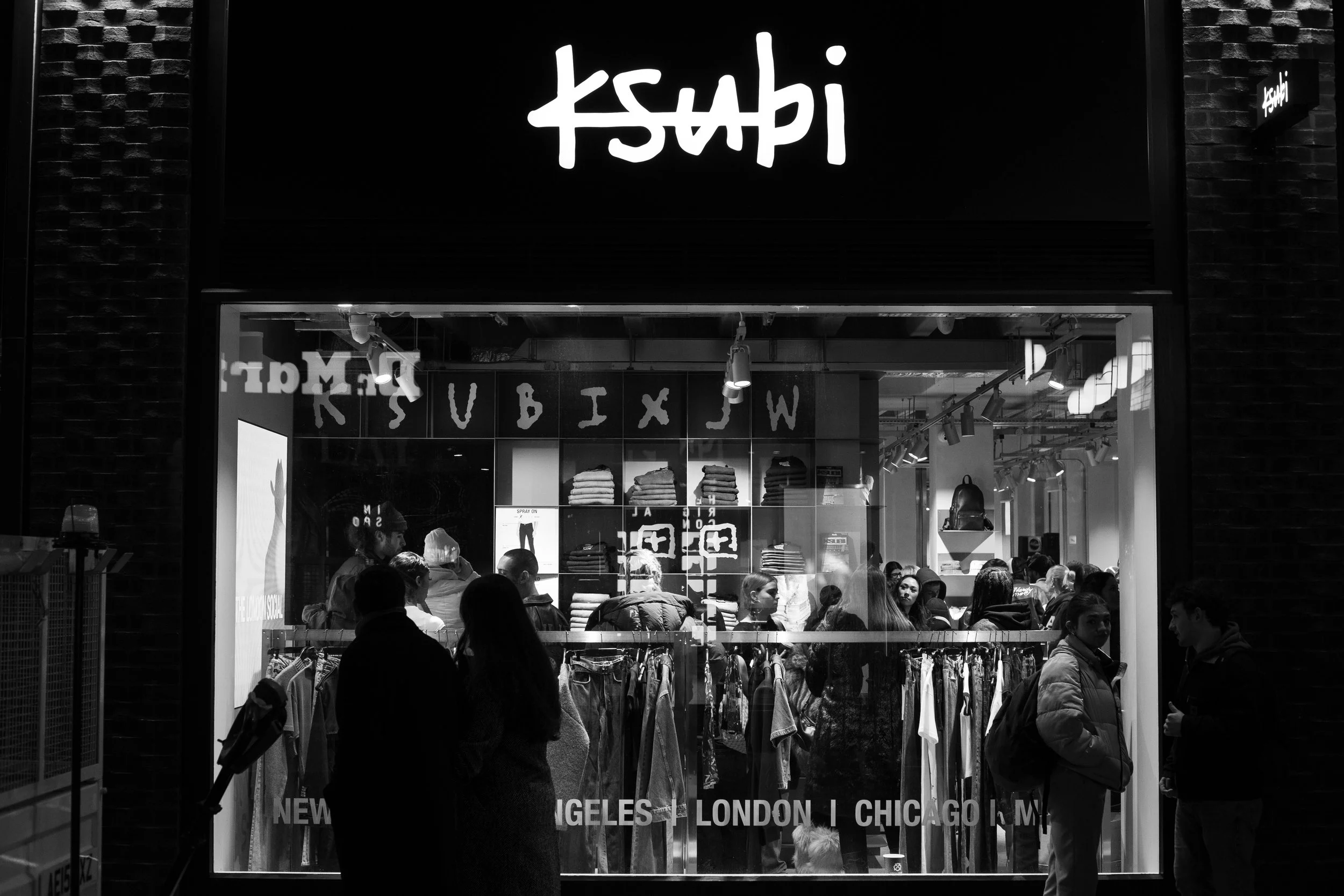 Night view of a clothing store window with people shopping inside and outside, featuring clothing racks and folded sweaters on display, with the store's sign above the window