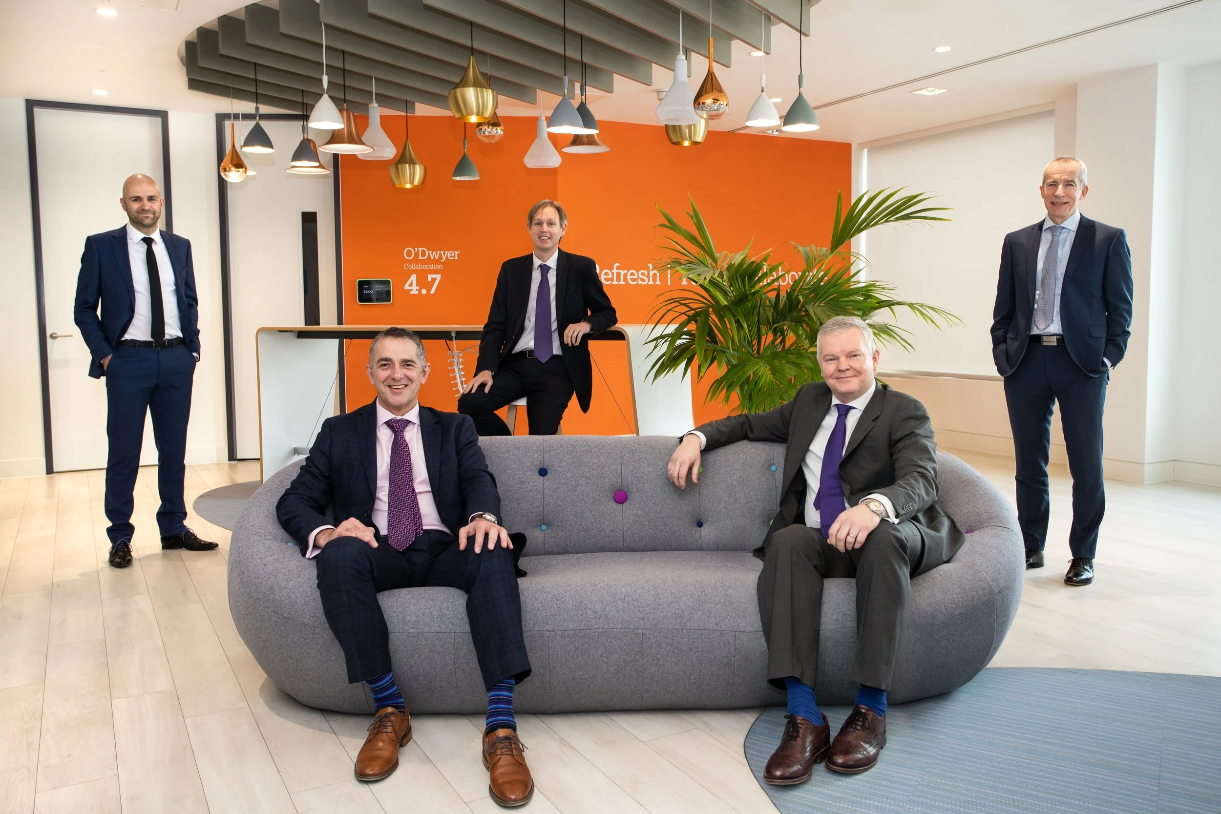 Five men in business suits in a modern office lobby, with two seated on a gray sofa, one standing to the left, one on the far right, and one leaning on a white desk in the background, with an orange wall and hanging light fixtures.