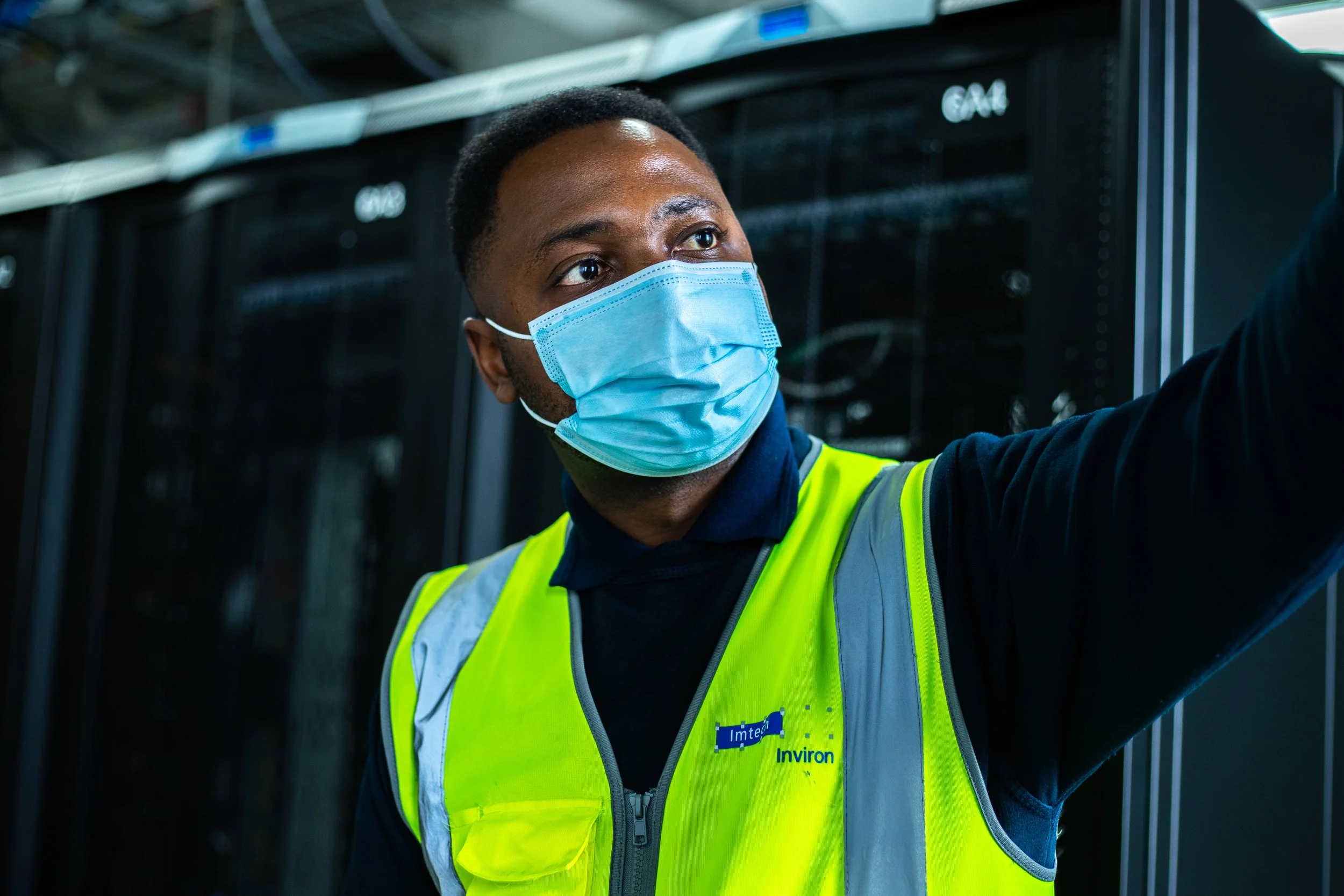 A man wearing a blue face mask and a reflective yellow safety vest standing in front of server racks.