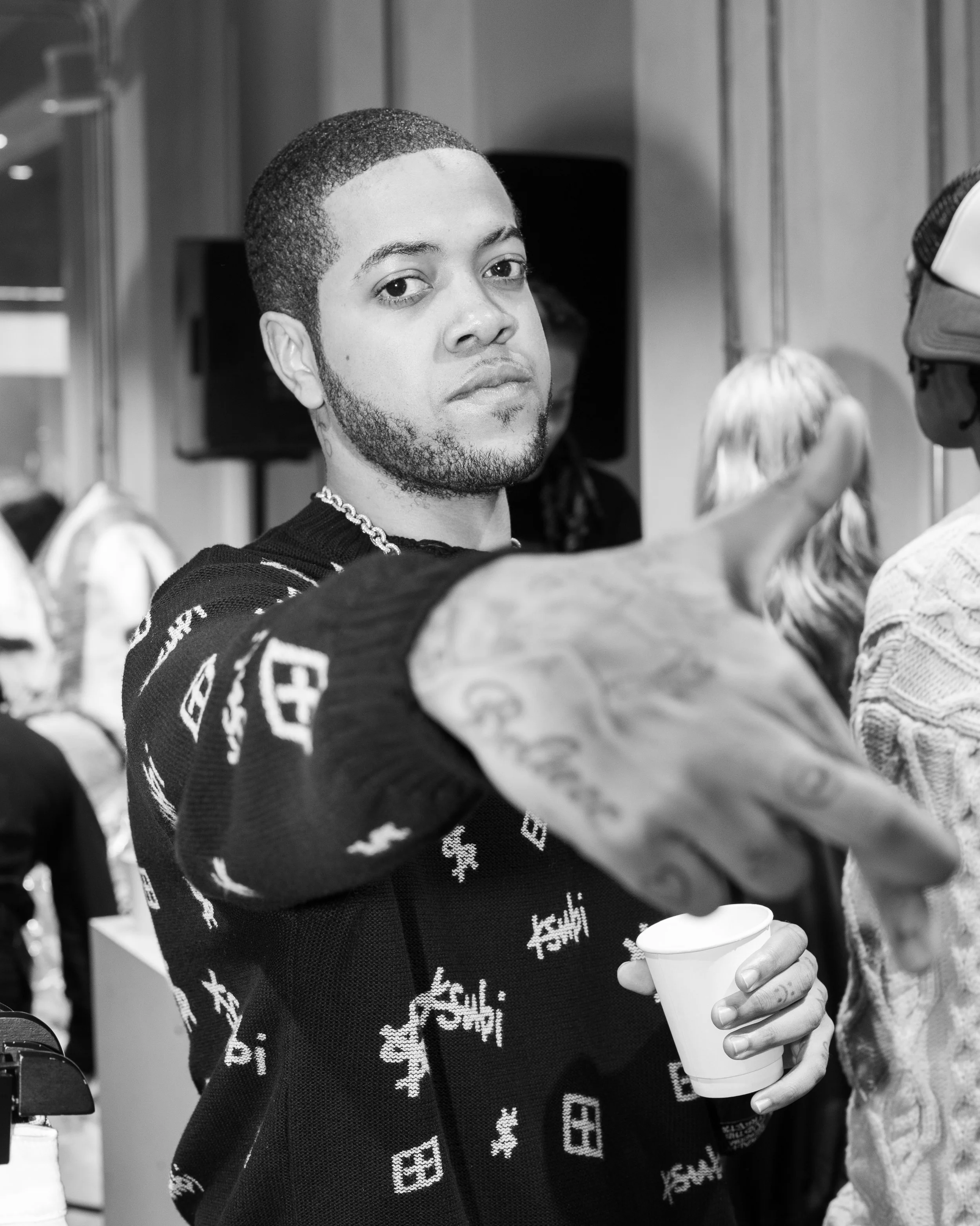 A young man with short hair and a beard looks at the camera while holding a cup and extending his hand toward the camera in a social setting.
