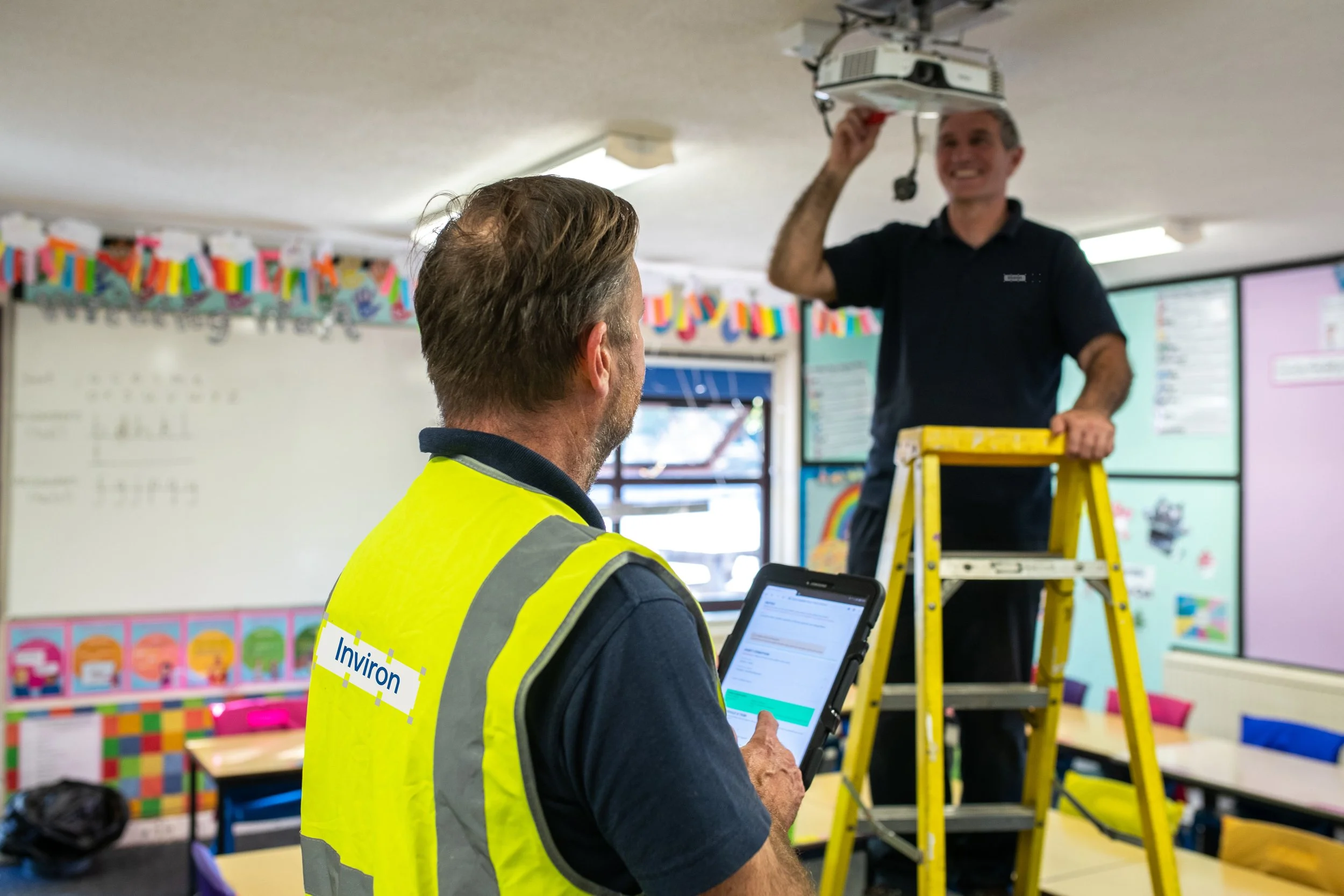 A man wearing a yellow safety vest labeled 'Inviron' is holding a tablet, looking at a second man who is on a ladder, working on a ceiling projector in a classroom decorated for a celebration.