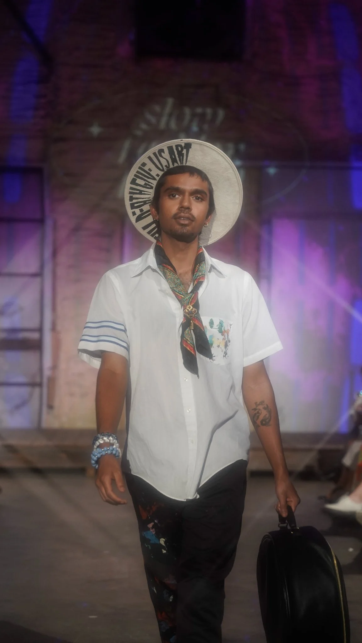 A man with short dark hair and a beard walking on a stage, wearing a white shirt, colorful scarf, bracelets, and a hat with text, carrying a black rounded briefcase, with colorful stage lighting and a brick wall background.