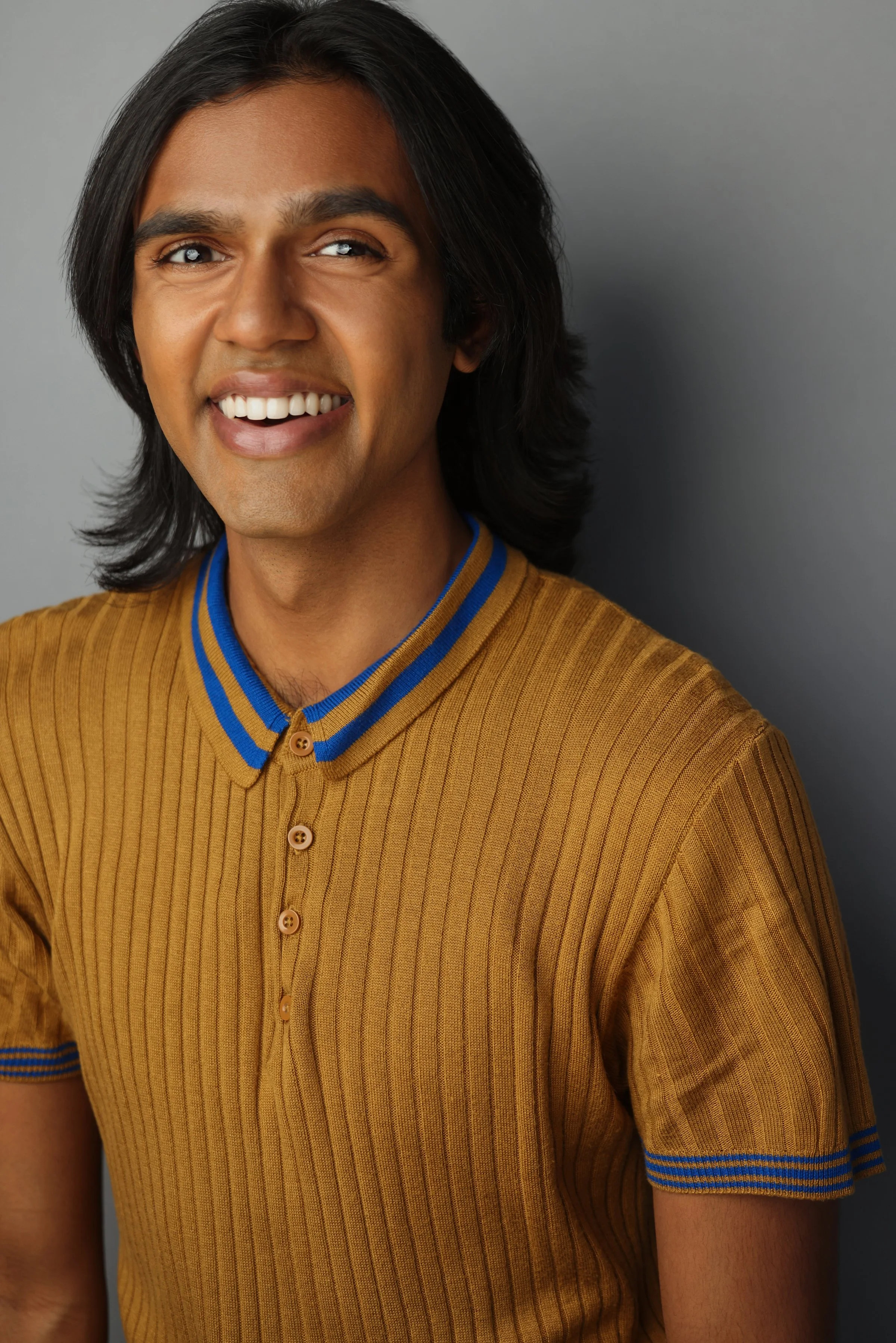 A man with long black hair, smiling, wearing a mustard yellow polo shirt with blue stripes on the collar and sleeves, standing against a gray background.