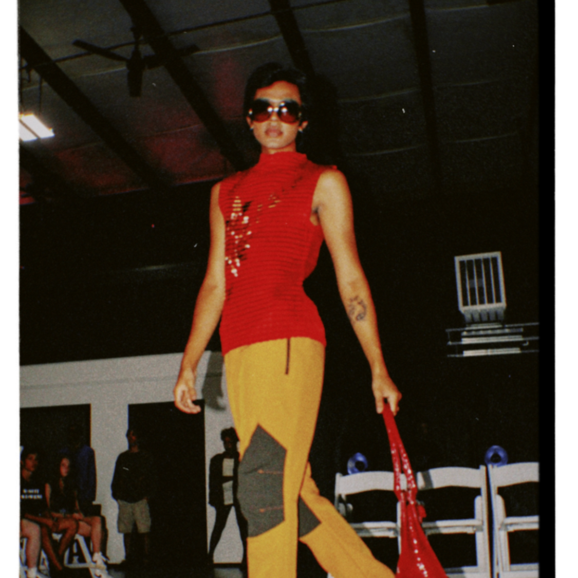 A woman walking on a runway during a fashion show, wearing a red sleeveless top with gold and black pants, sunglasses, and carrying a red bag.