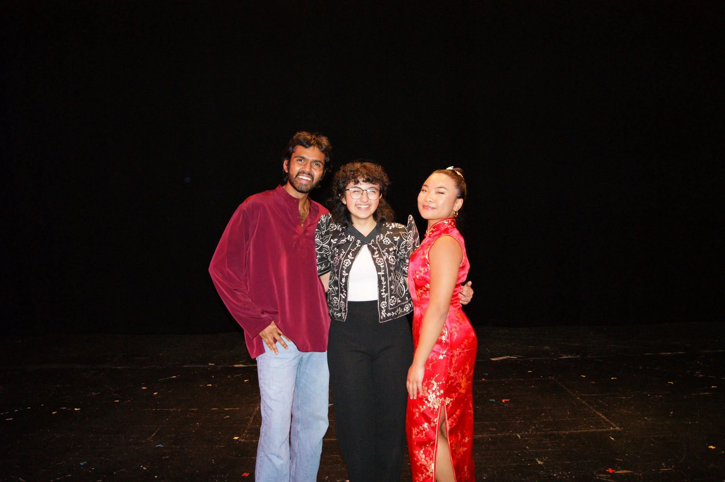 Three people smiling and posing together on stage with a black background, two women and one man, dressed in colorful attire.