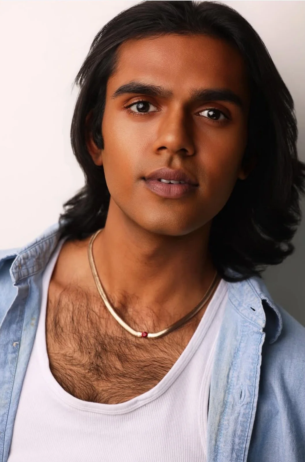 Close-up of a young man with medium skin tone, long dark hair, wearing a white tank top, a light denim shirt, and a silver necklace with a small red bead, looking directly at the camera.