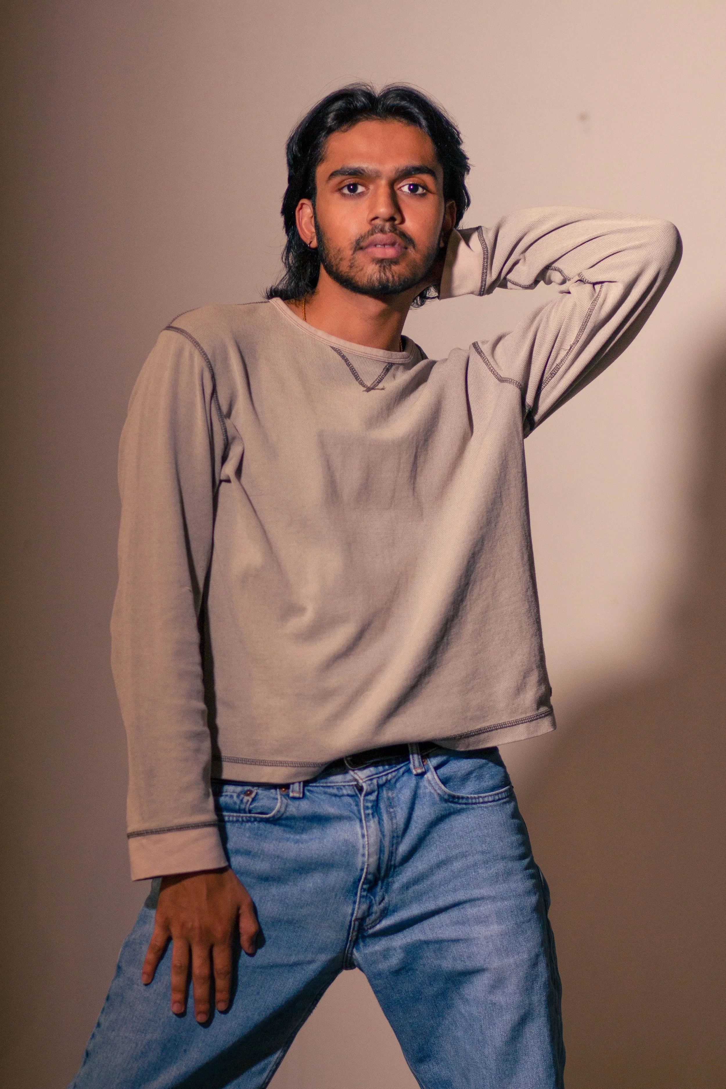 A young man with dark hair, mustache, and beard wearing a beige long-sleeve shirt and blue jeans posing with one hand behind his head against a plain wall.