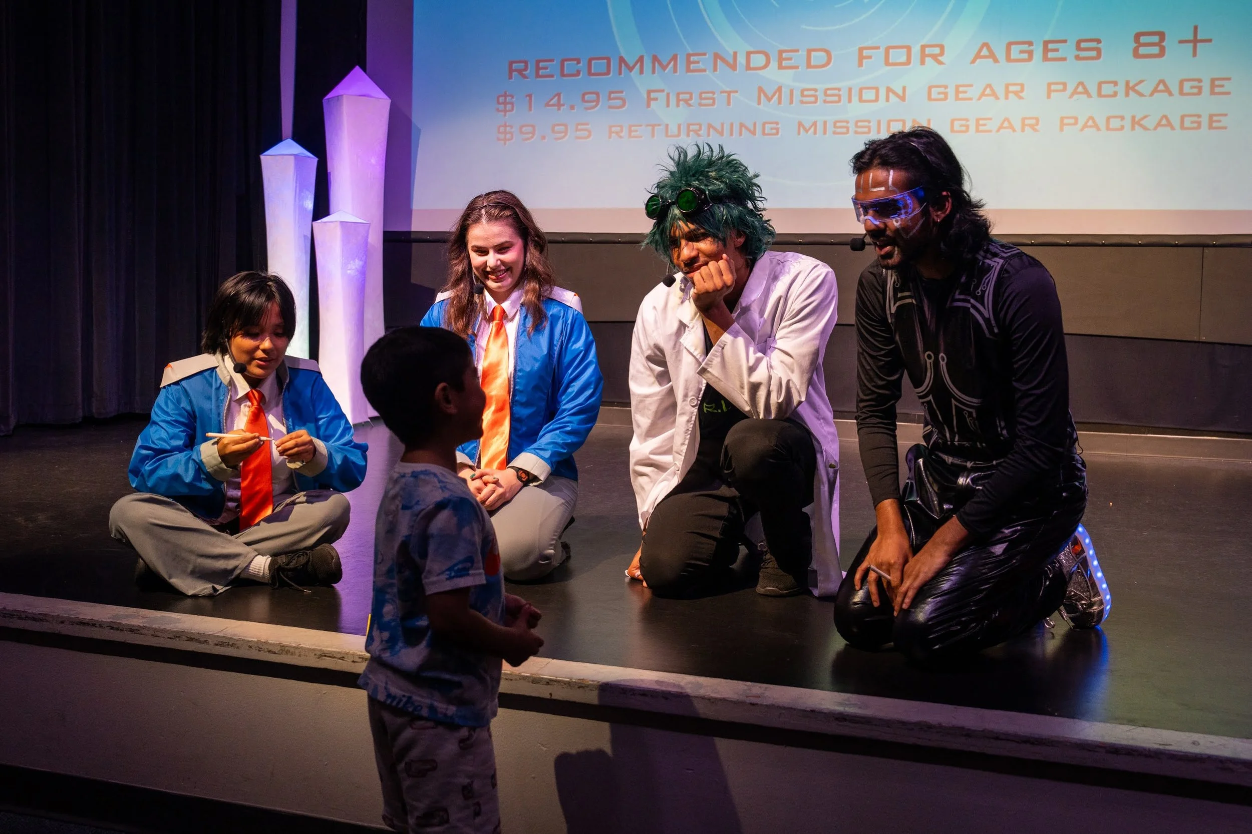Group of children and adults dressed as scientists and space characters, sitting and kneeling on a stage with a projected space-themed background and text about recommended age and gear package.