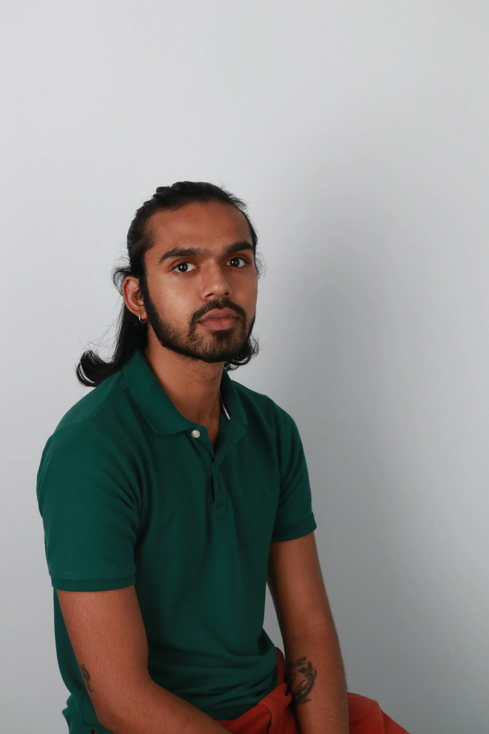 Portrait of a man with long dark hair and a beard, wearing a green polo shirt and sitting against a plain gray wall.