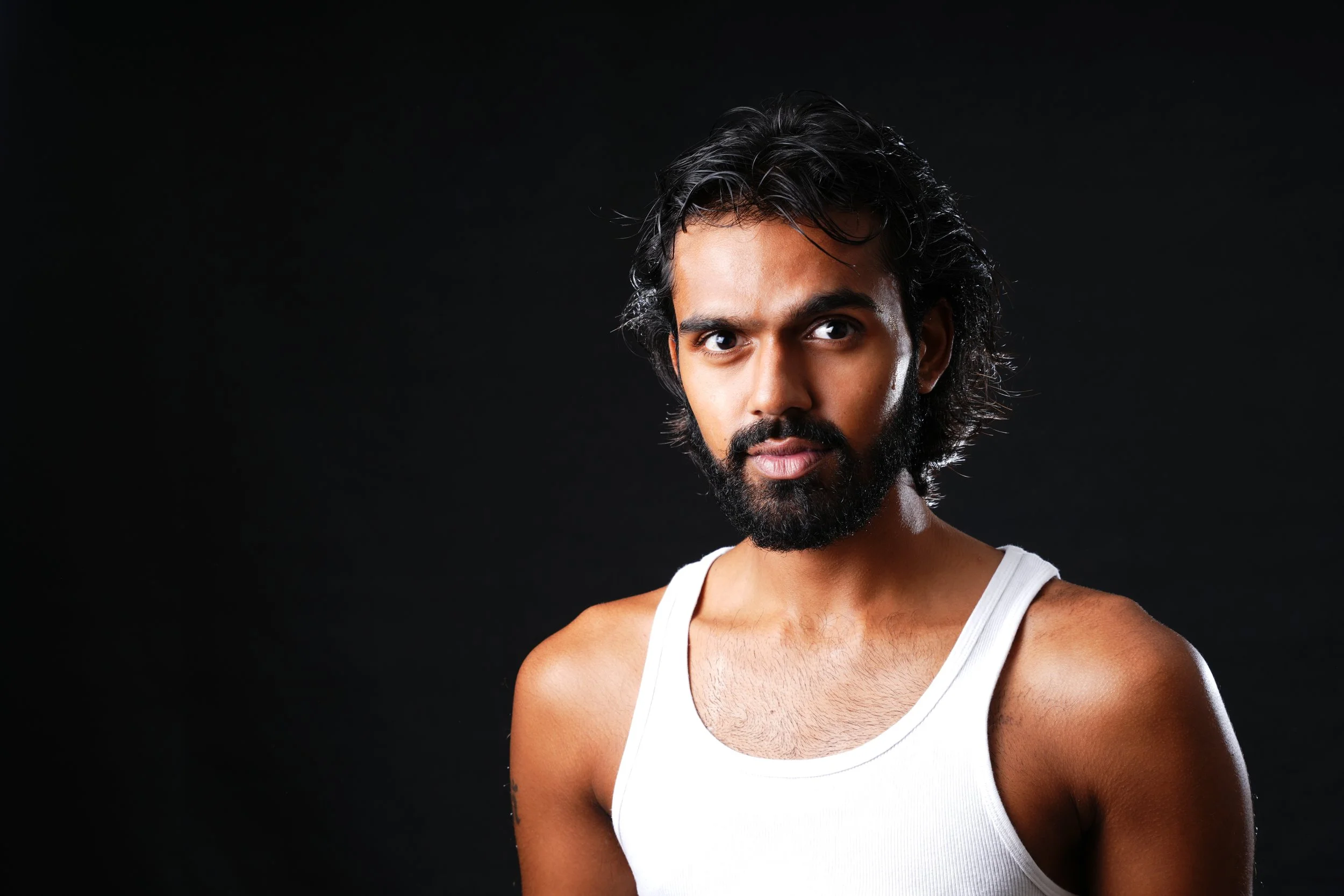 Portrait of a man with dark hair, a beard, and wearing a white tank top against a black background.