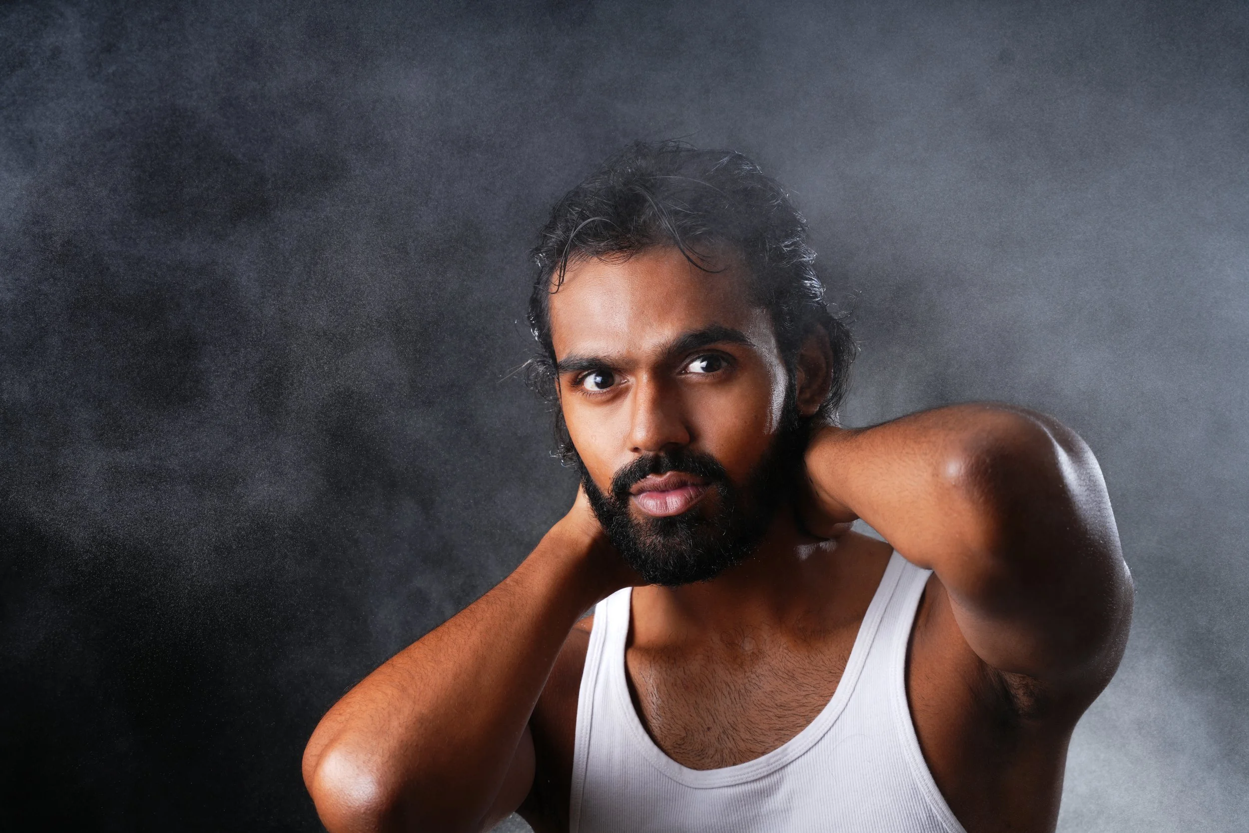 A man with dark hair, beard, and mustache in a white tank top, looking directly at the camera with hands behind his neck against a dark, smoky background.