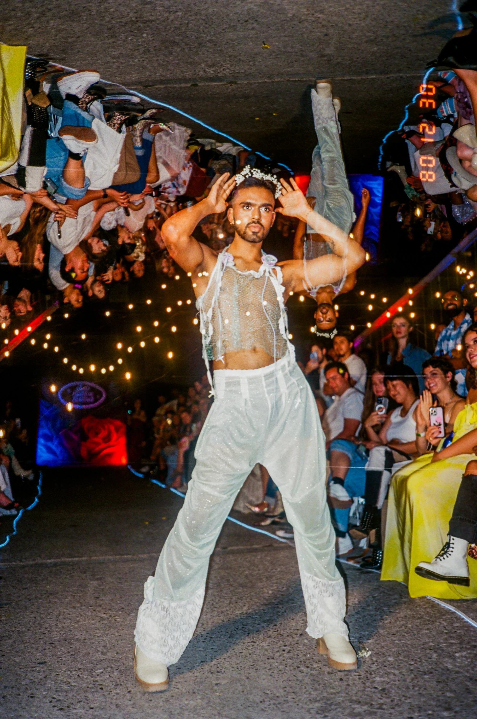 A man walking down a runway at a fashion show wearing loose white pants with lace details and a sheer, sleeveless top, while an audience watches and takes photos.