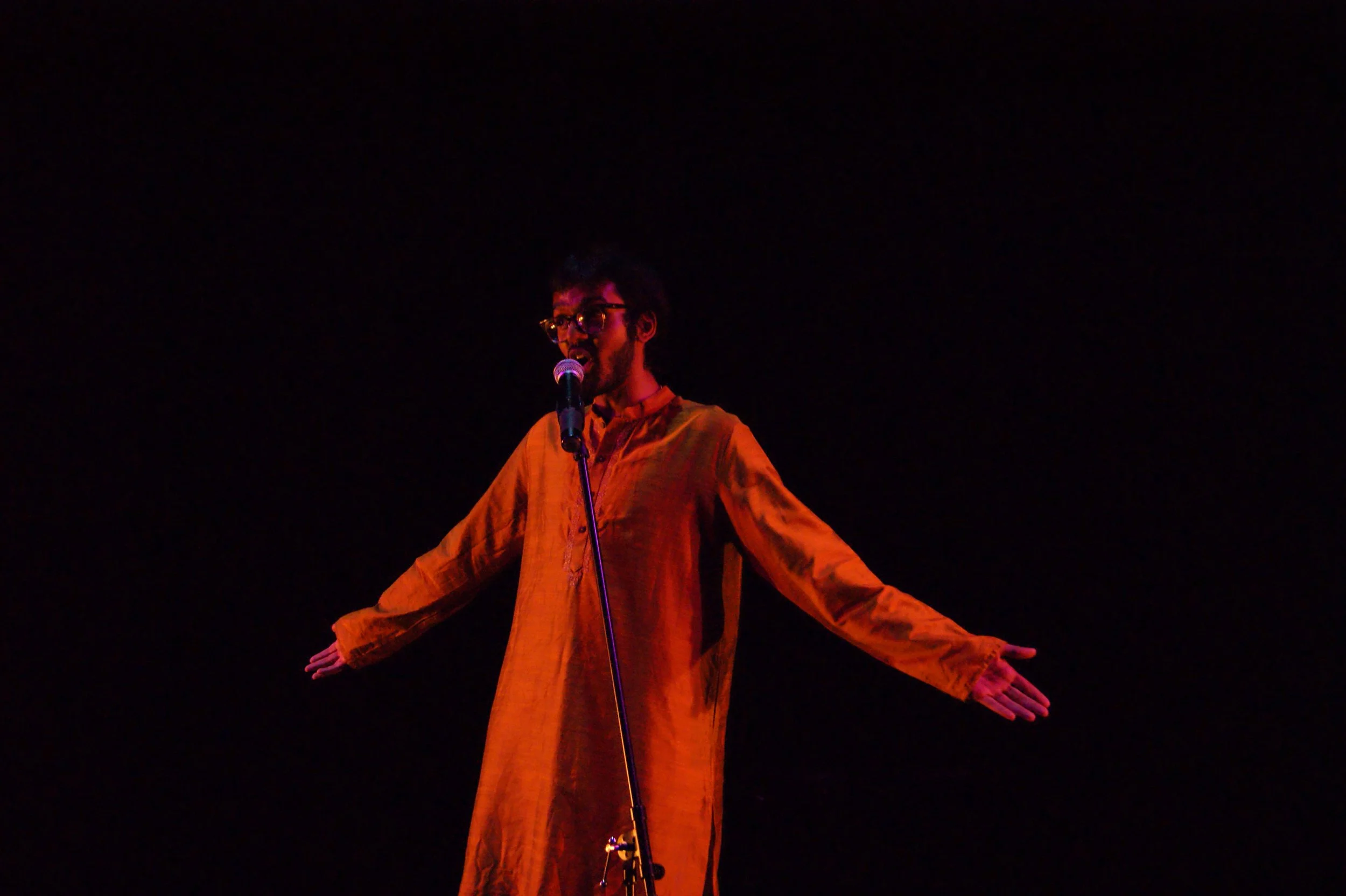 A person standing on a stage with arms outstretched, wearing an orange outfit and speaking into a microphone against a black background.