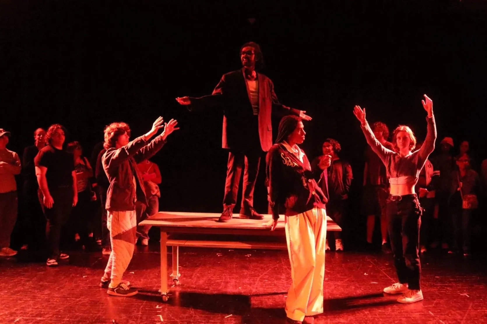 A theatrical performance with a woman on a table center stage, dressed in dark clothing, with her arms outstretched. A person with short hair and a patterned outfit stands in front of the table. Four young women around them, some raising their hands, appear to be participating in a dramatic scene, with other onlookers in the background. The scene is lit with red lighting, creating a dark, intense atmosphere.
