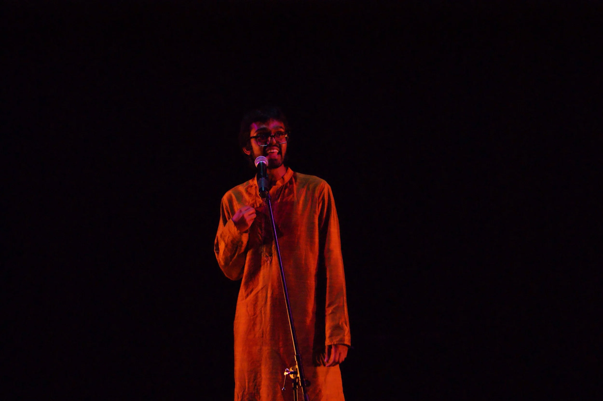 A man wearing glasses and an orange traditional outfit speaking into a microphone against a dark background.