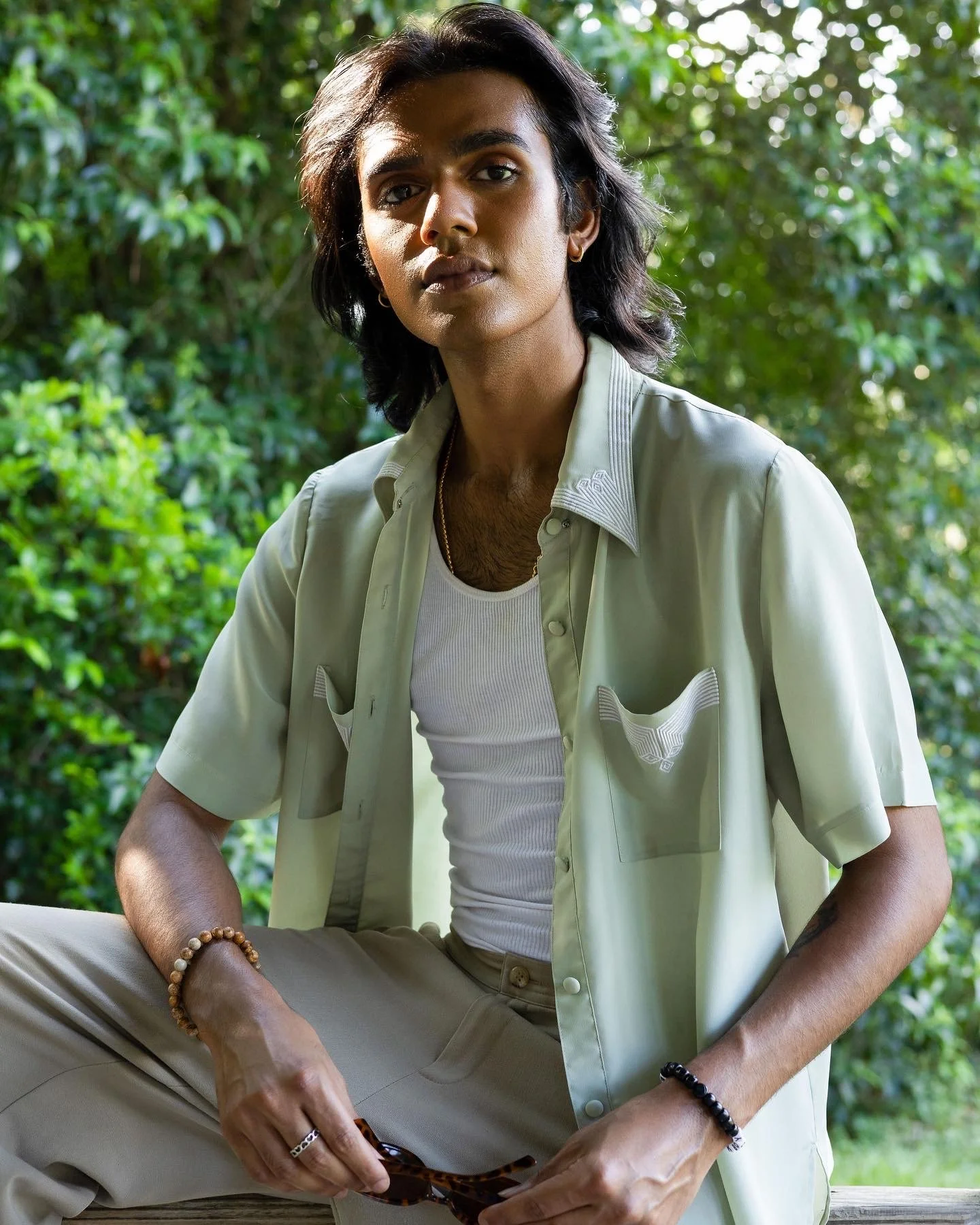 A young person with long dark hair sits outdoors on a bench, wearing a light-colored short-sleeved shirt over a white tank top, beige pants, and bracelets. They are holding sunglasses and looking into the camera with a serious expression, with green 