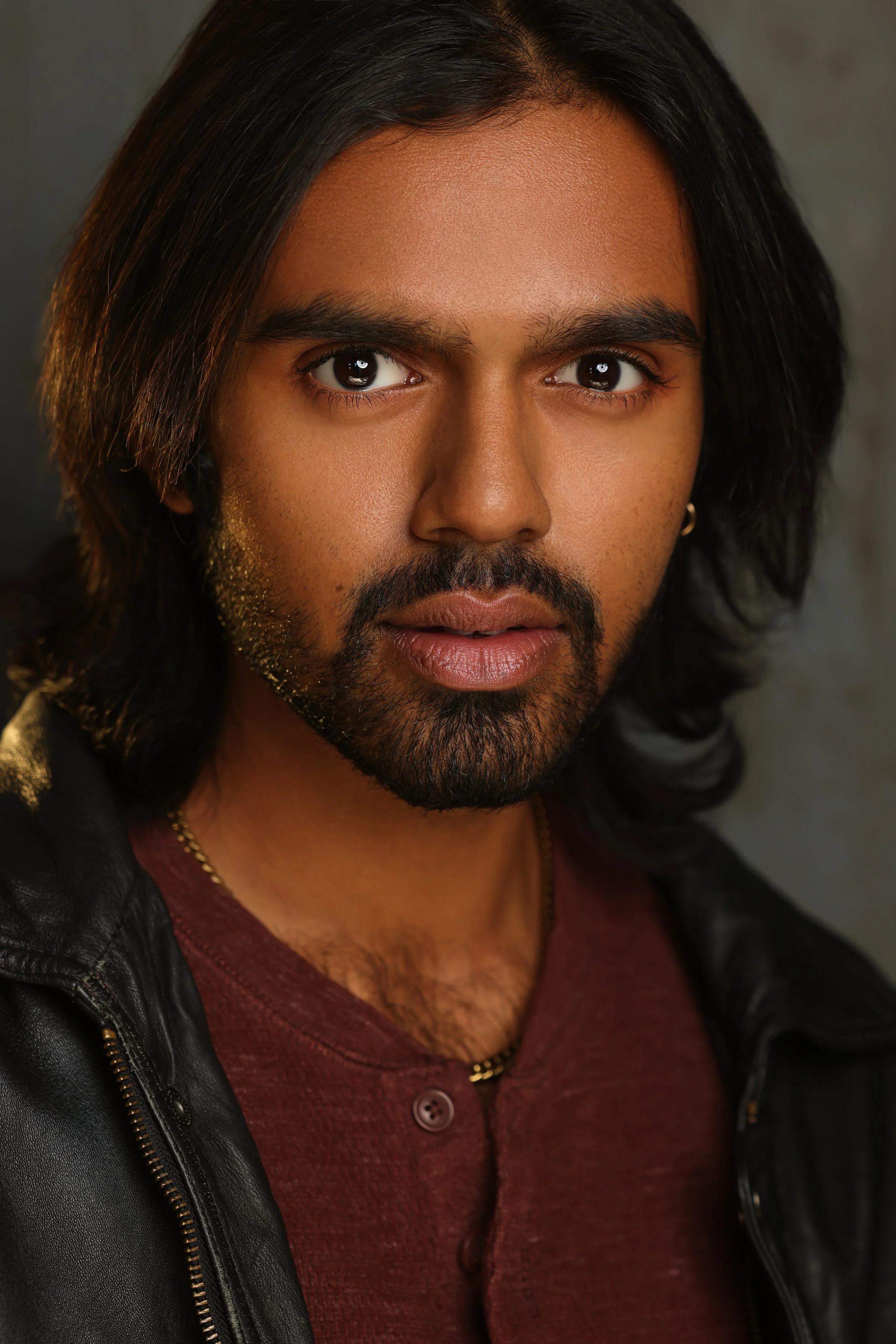Close-up portrait of a man with long black hair, dark eyes, light brown skin, a beard, wearing a leather jacket over a red shirt, with a gold chain.