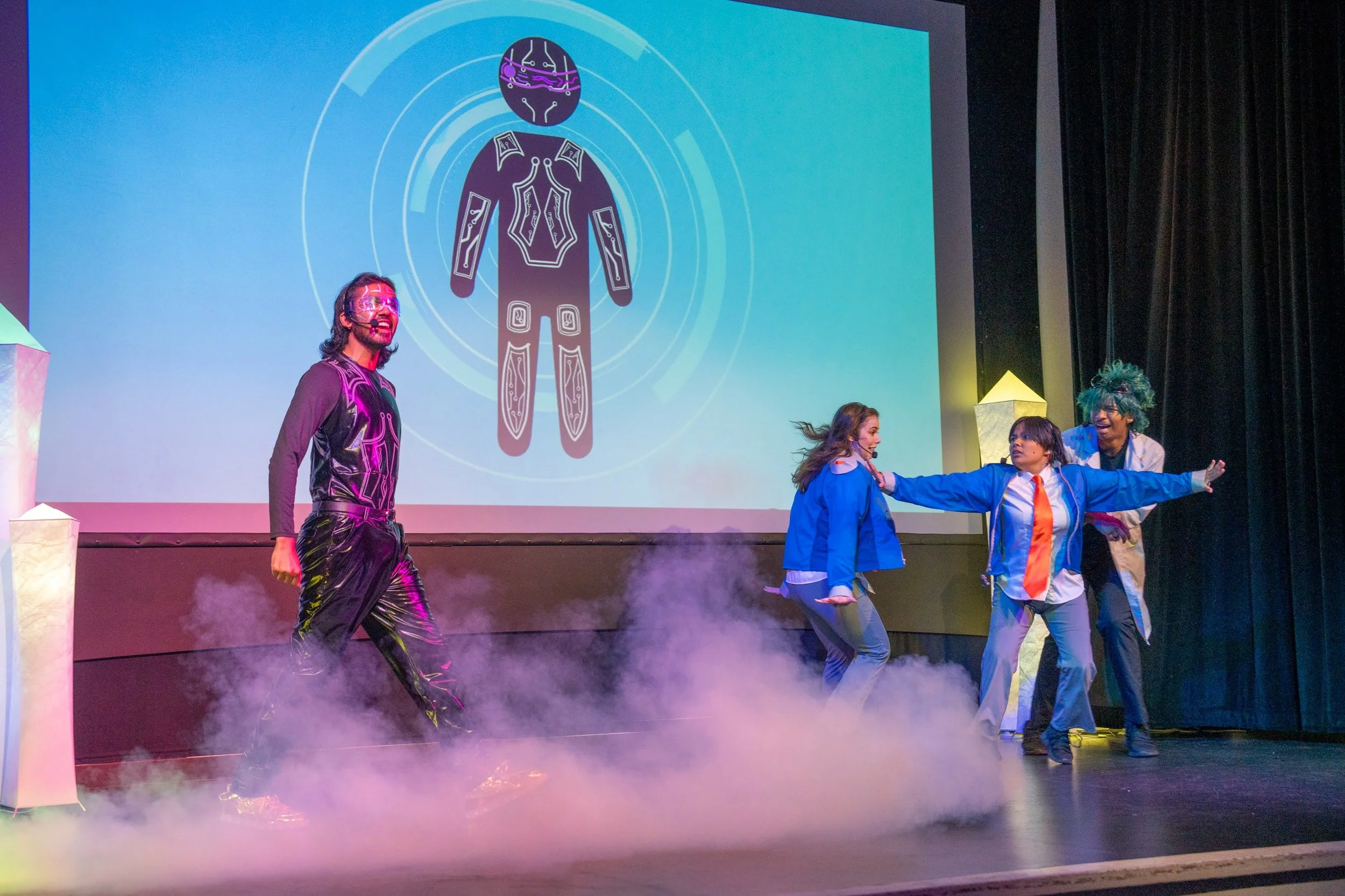 Performers on stage during a science fiction-themed play or presentation, with a large digital diagram of a human body projected on screen behind them. One performer is dressed in a black futuristic costume, and three others are in colorful costumes, engaging in a theatrical scene with fog effects.