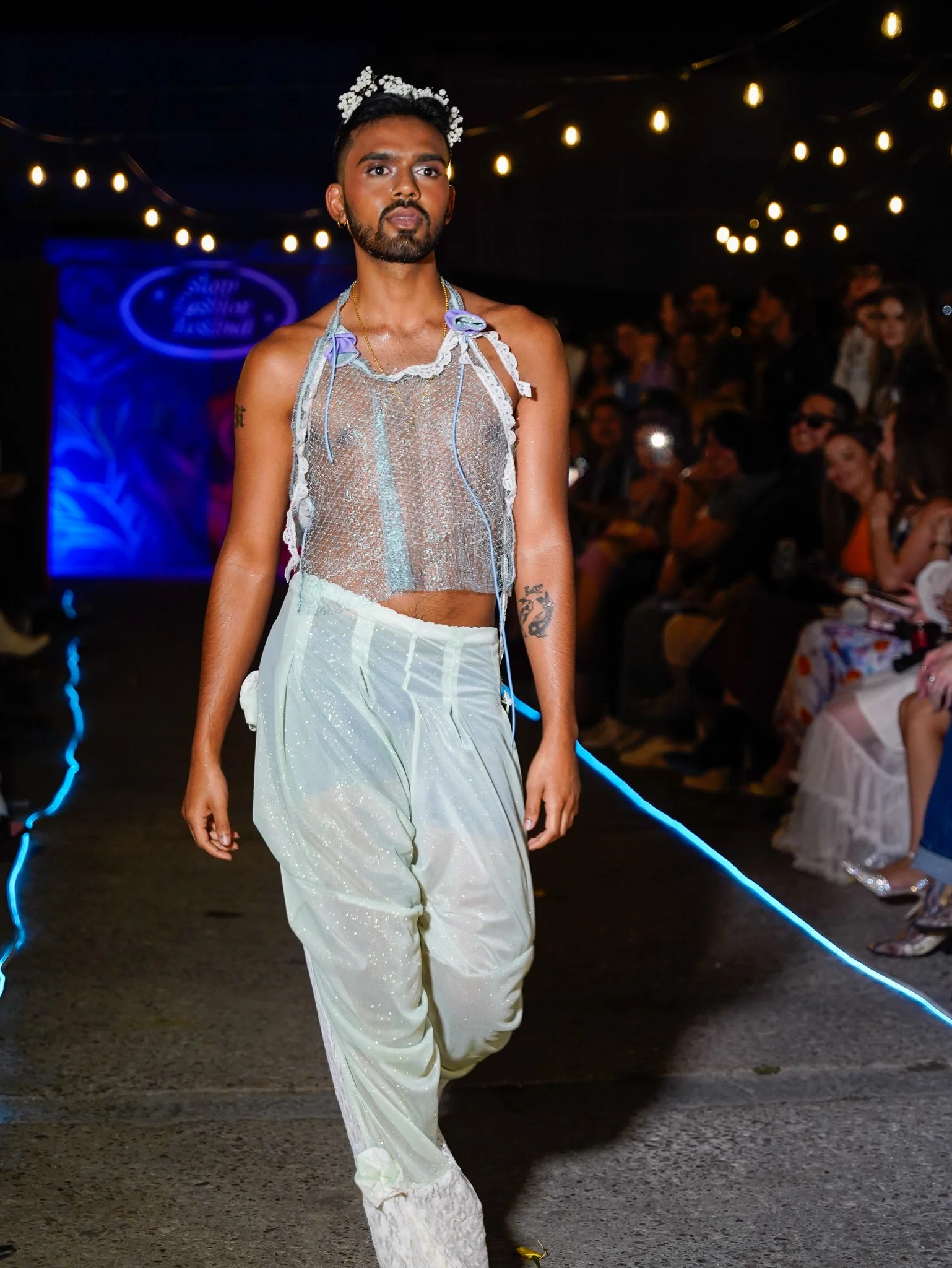 A male model walking down a runway during a fashion show, wearing sheer top, loose white pants, and a floral headpiece, with audience members watching in the background.