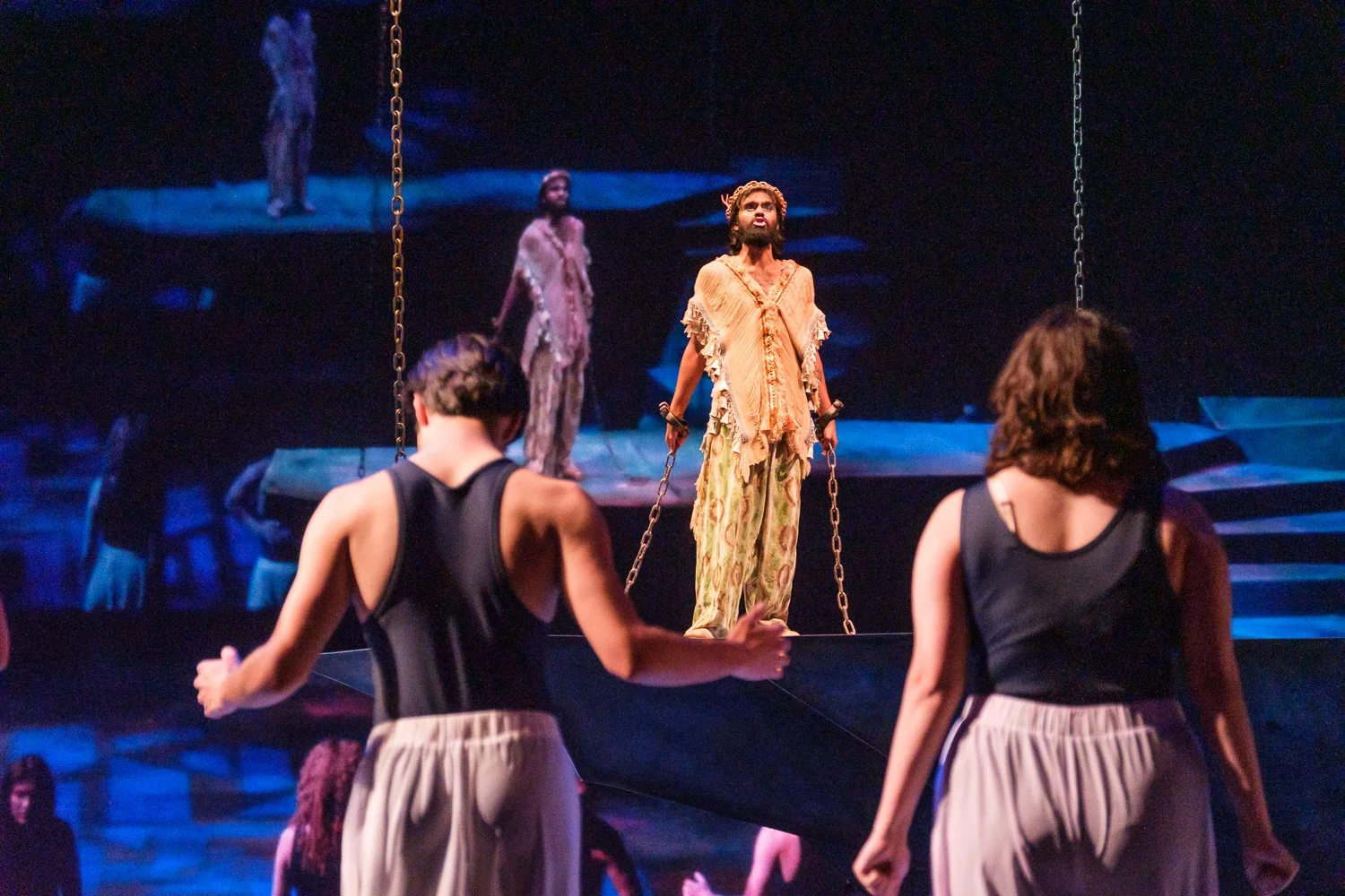 Stage scene from a theatrical performance with multiple actors. One actor, dressed in tattered clothing, stands center with chains hanging from their wrists. Two women in black tops and beige skirts are in foreground, performing or preparing for a scene. The background features a dark set with blue lighting and distant figures.