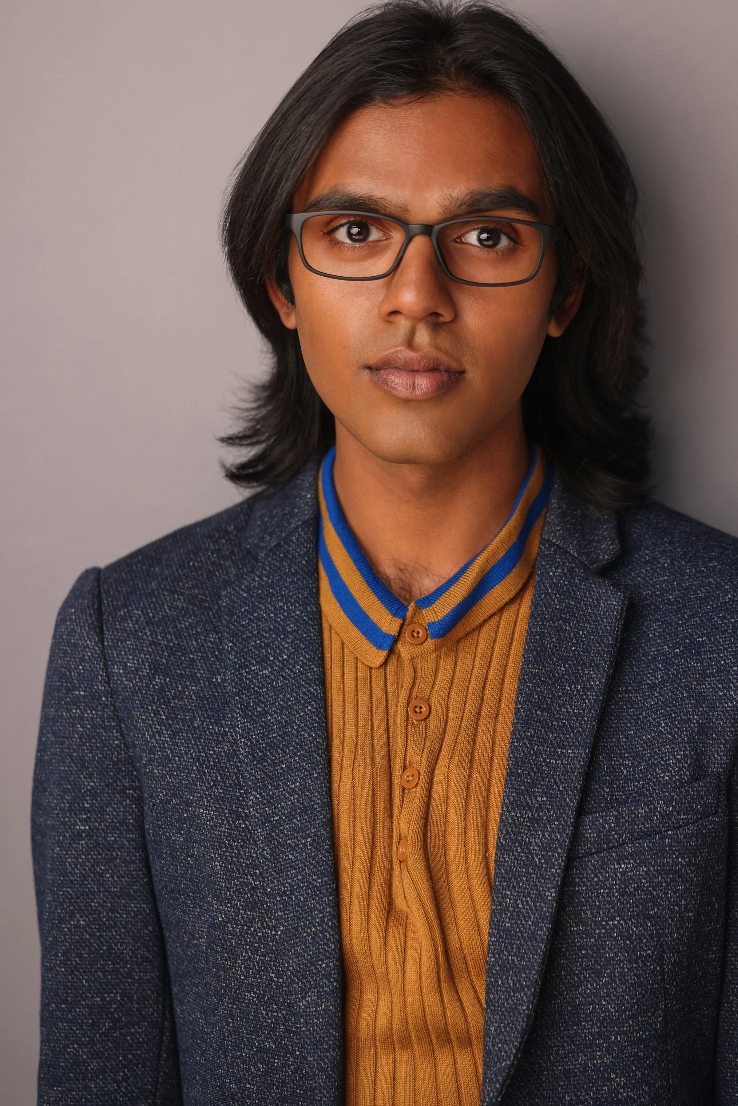 Portrait of a young man with dark, shoulder-length hair, wearing glasses, a mustard yellow ribbed shirt with a blue striped collar, and a dark blue blazer, standing against a gray background.