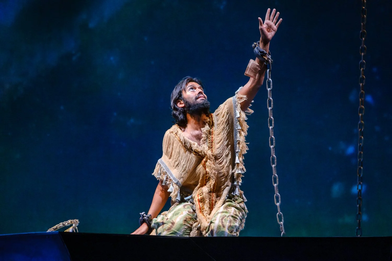 A man with long dark hair and a beard, wearing a torn, earthy-toned costume, kneels on a platform and reaches up towards the sky, holding onto a chain, against a starry night background.