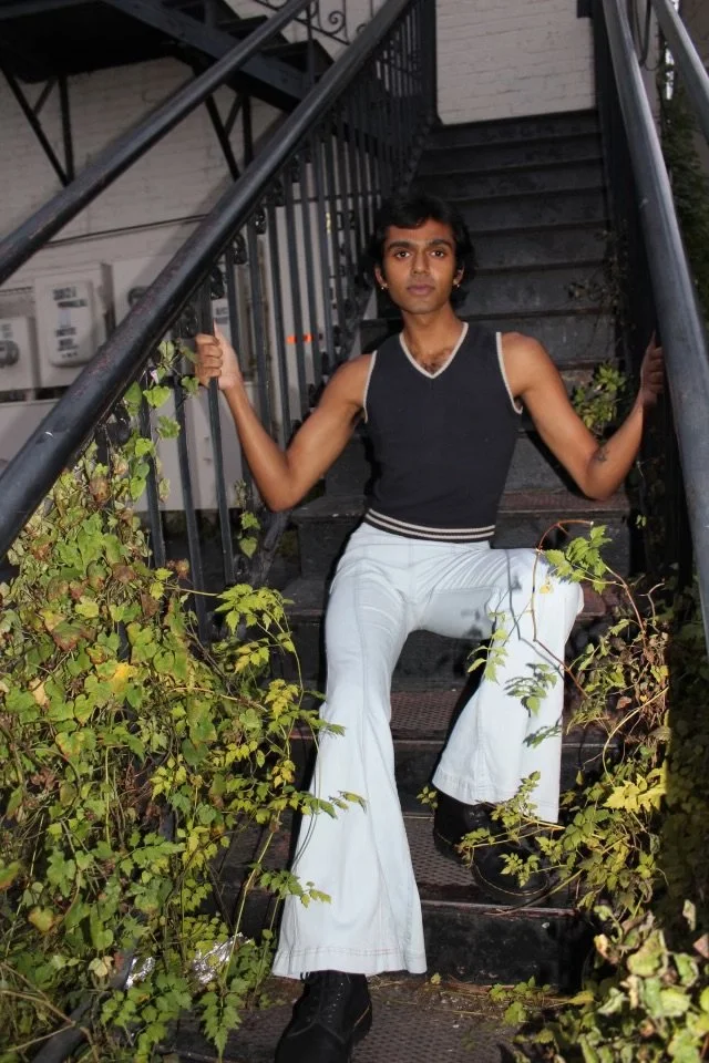 A young person with dark hair sitting on outdoor metal stairs, holding onto the railing, surrounded by green plants, wearing a sleeveless black shirt with white trim, white pants, and black shoes.