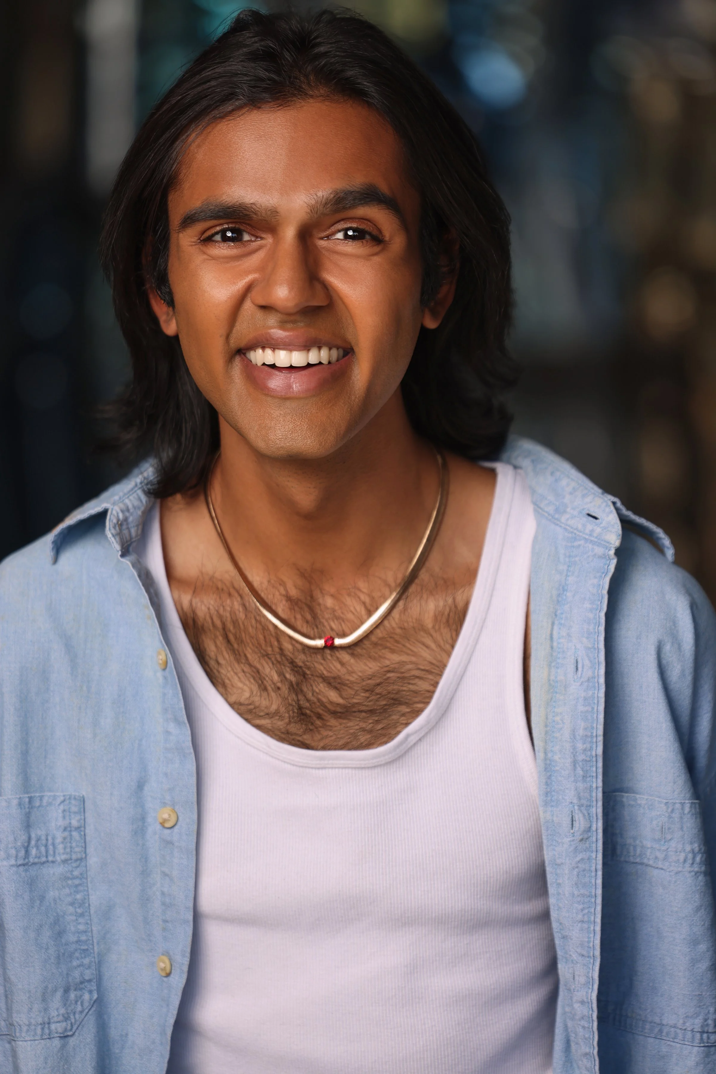 A smiling man with shoulder-length dark hair, wearing a denim shirt over a white tank top, with a thin necklace, standing outdoors with blurred background.