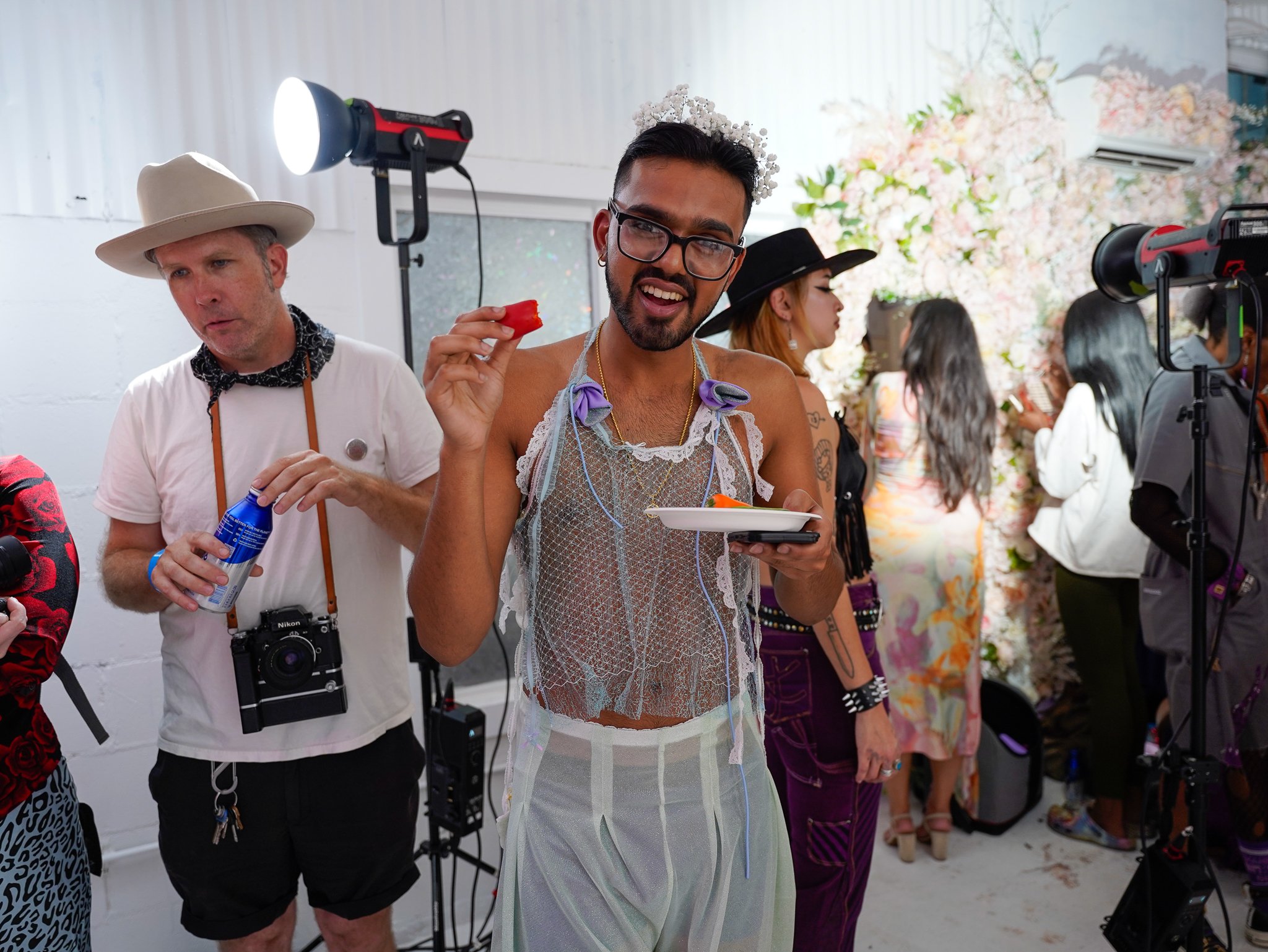 People at a party or event, with a man in the center holding a plate of food and a piece of food in his hand, wearing a sheer top, and others in the background in stylish outfits, decorated with flowers.