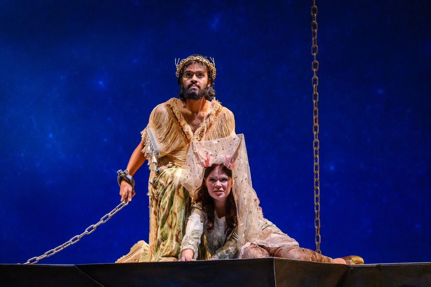 Two actors on stage, one standing and one kneeling, with a dark blue starry background and hanging chains, suggesting a theatrical performance.
