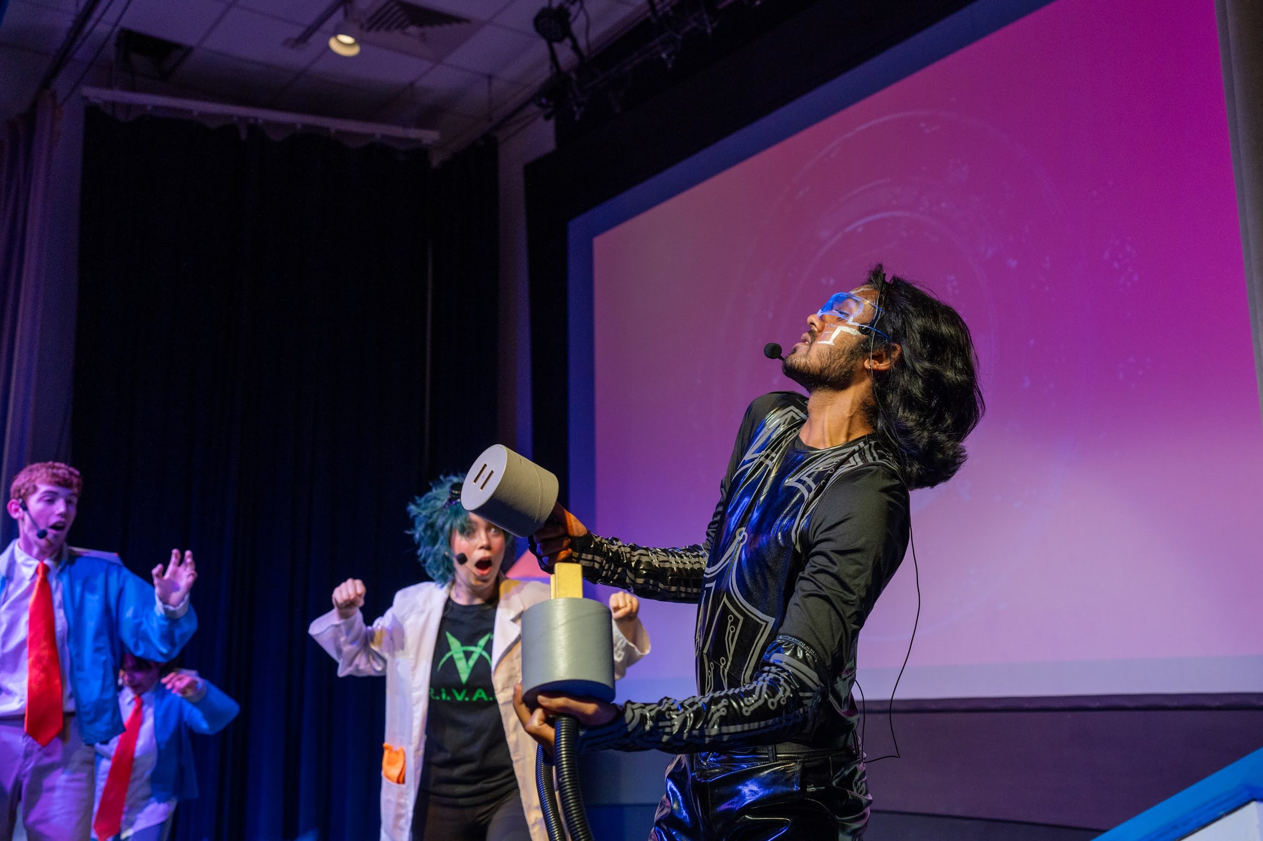 Performer with glasses and long black hair holding a rocket prop on stage with children reacting excitedly in the background.