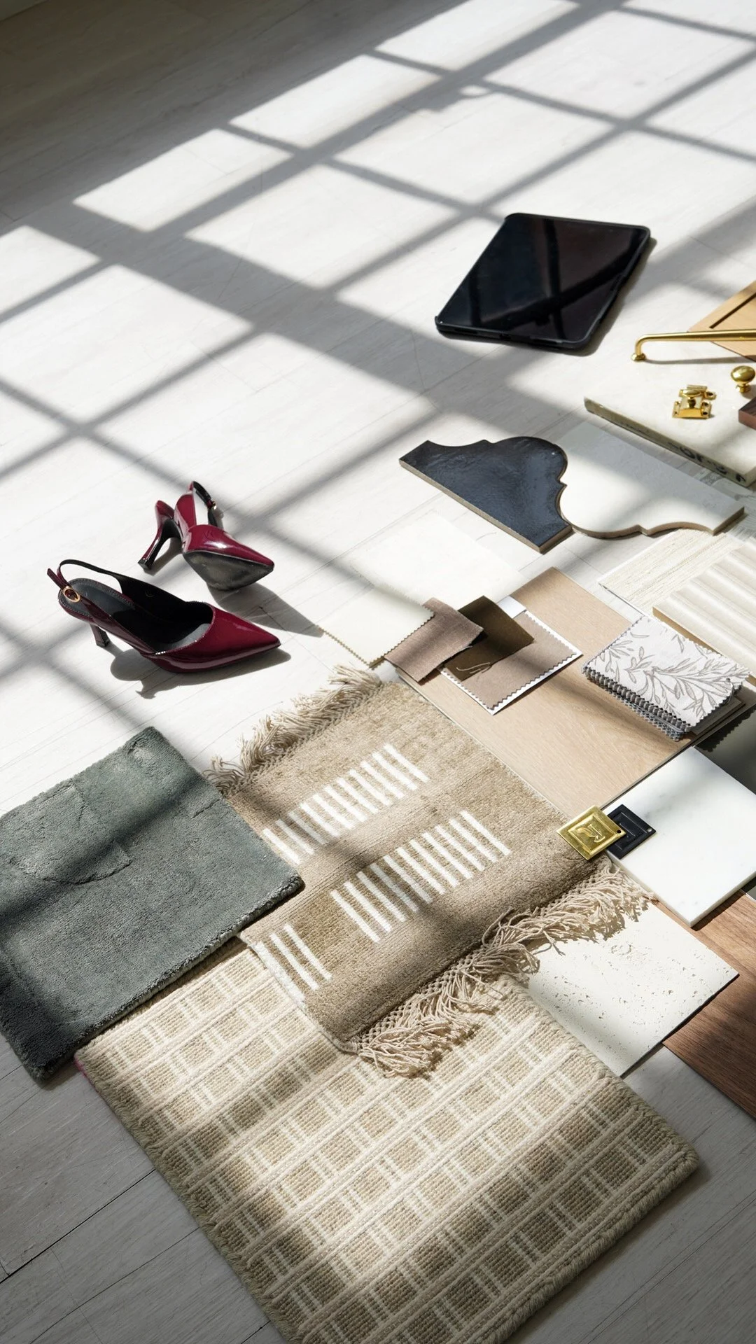 Samples of interior design materials including fabric swatches, rugs, shoes, a tablet, and decorative items arranged on a white wooden floor with sunlight and shadow patterns.