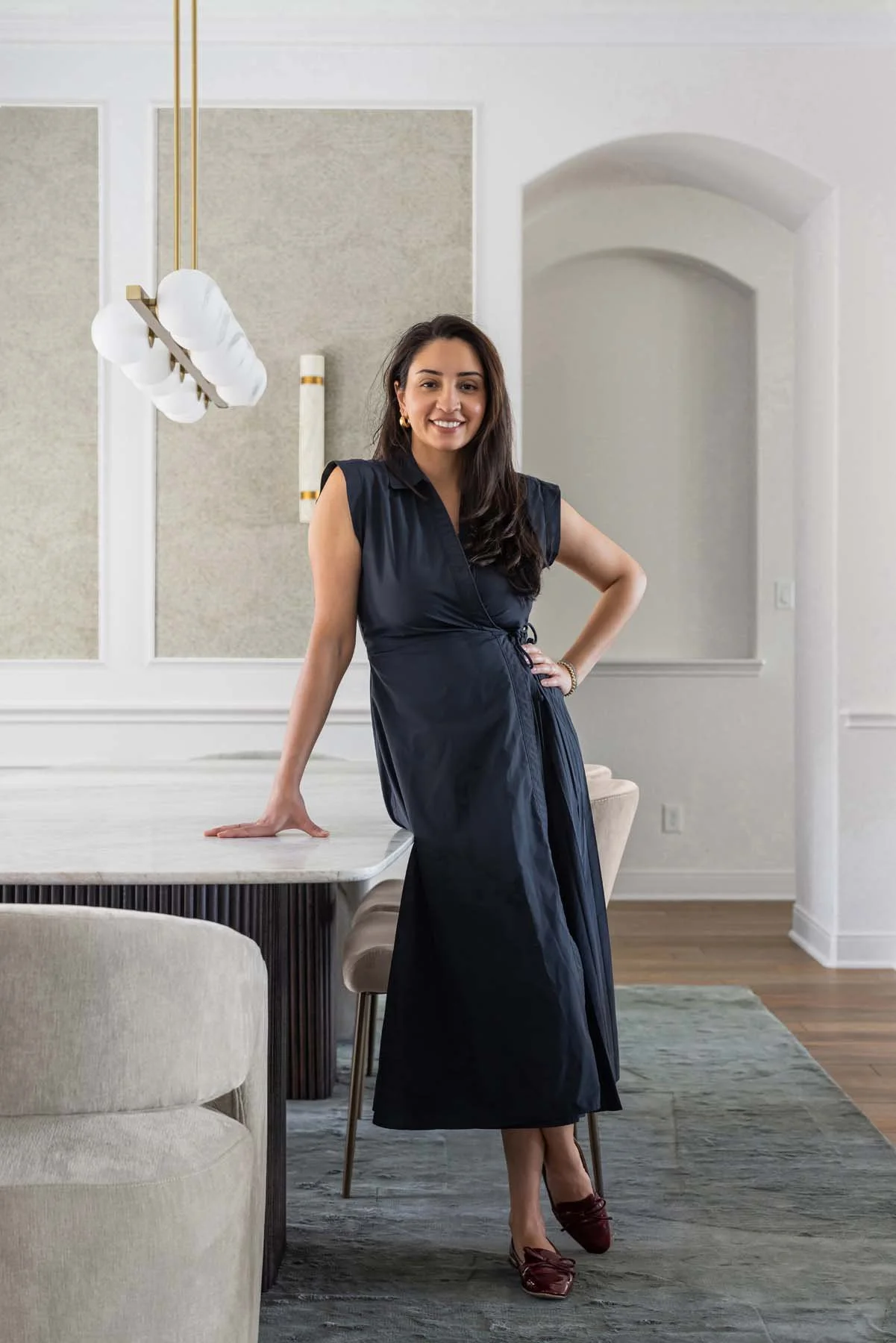 A woman in a black dress standing in a modern dining room, leaning against a marble table.