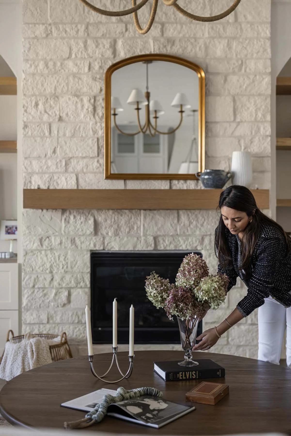 A woman arranging a bouquet of dried hydrangeas in a glass vase on a wooden table in a living room with a stone fireplace and a mirror above it.
