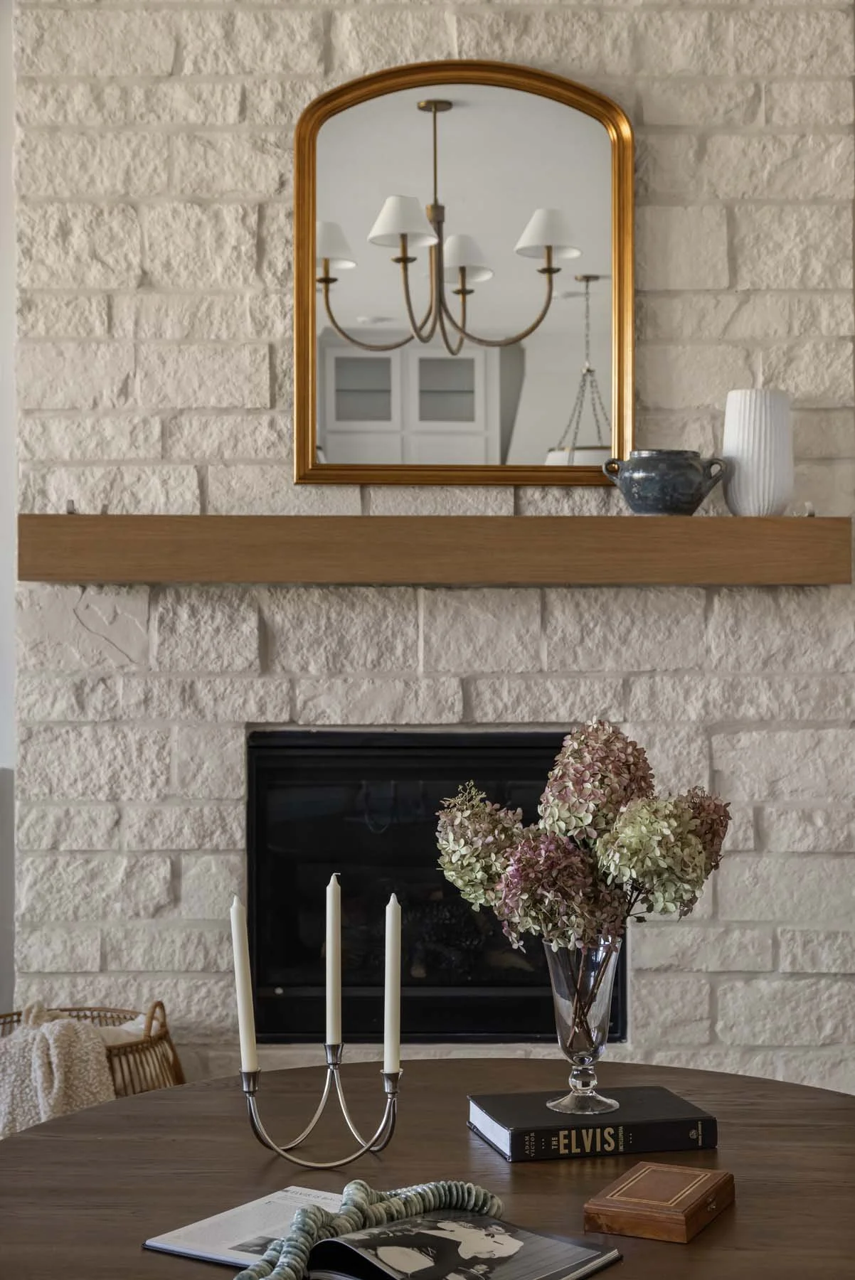 A living room with a stone fireplace, a wooden mantel with decorative vases, a mirror reflecting a chandelier, a wooden table with a candle holder, a book titled 'Elvis', a glass vase with dried hydrangeas, and an open magazine.