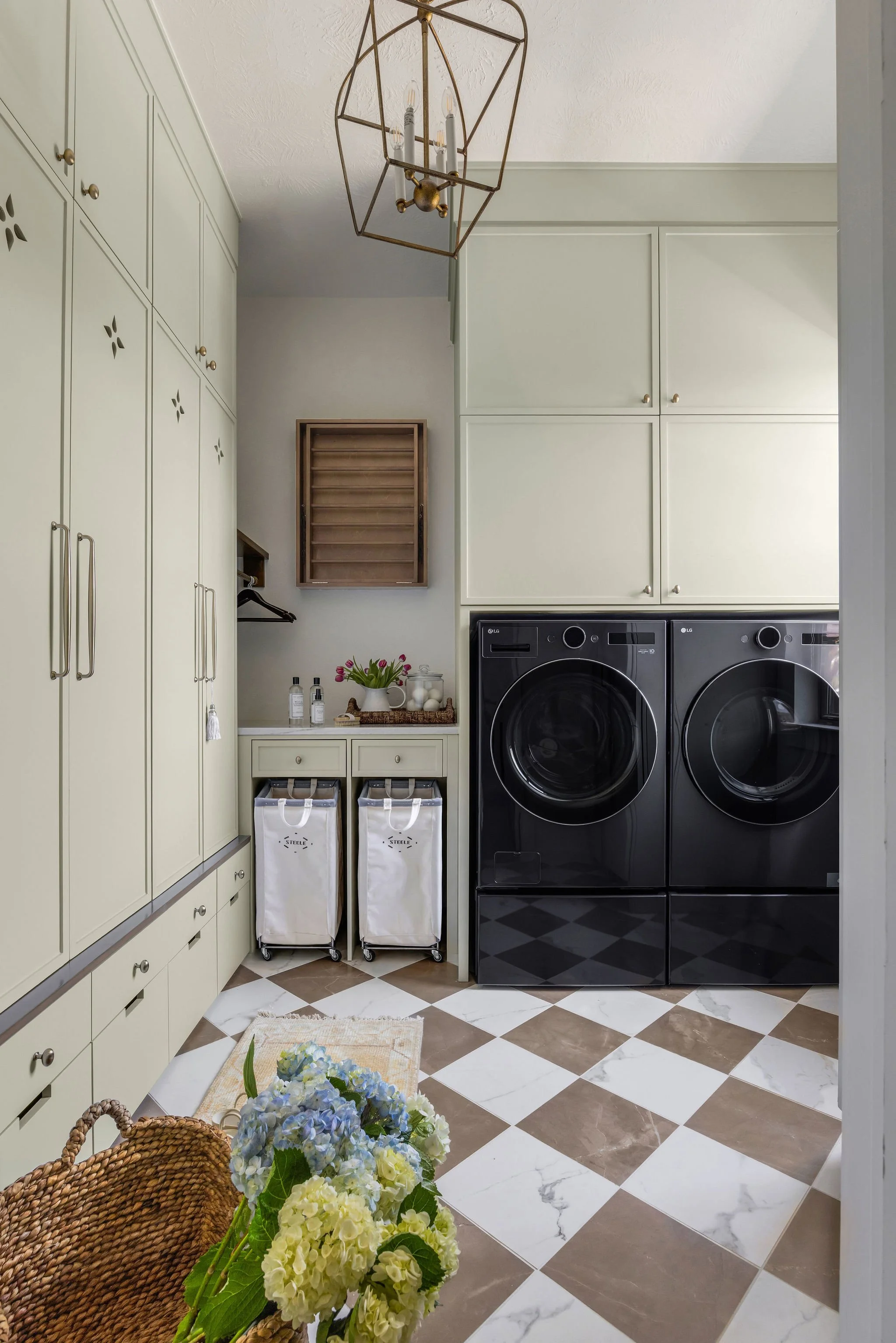 A laundry room with cream-colored cabinets, a black washing machine and dryer, a small table with flowers, plants, and bottles, and a patterned tile floor.